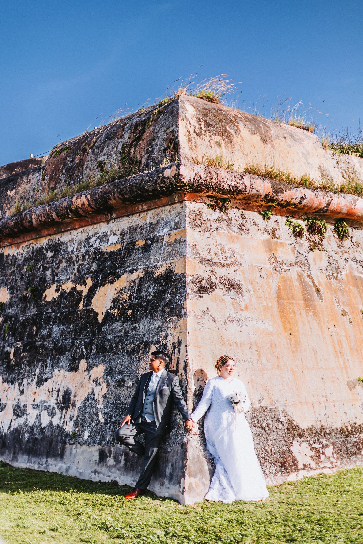 A wedding couple poses elegantly against a large rustic stone fortress wall.