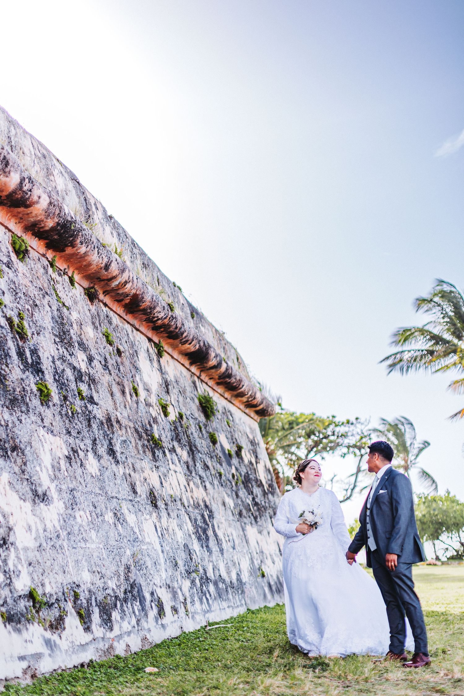 A bride and groom stand beside a towering weathered wall with palm trees in the background.
