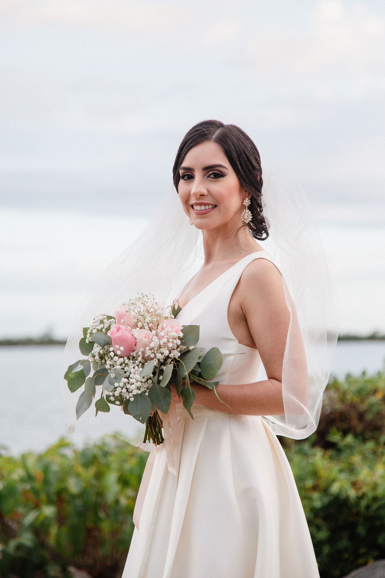 Smiling bride in white gown holding pink and white bouquet with veil, posing outdoors near water.