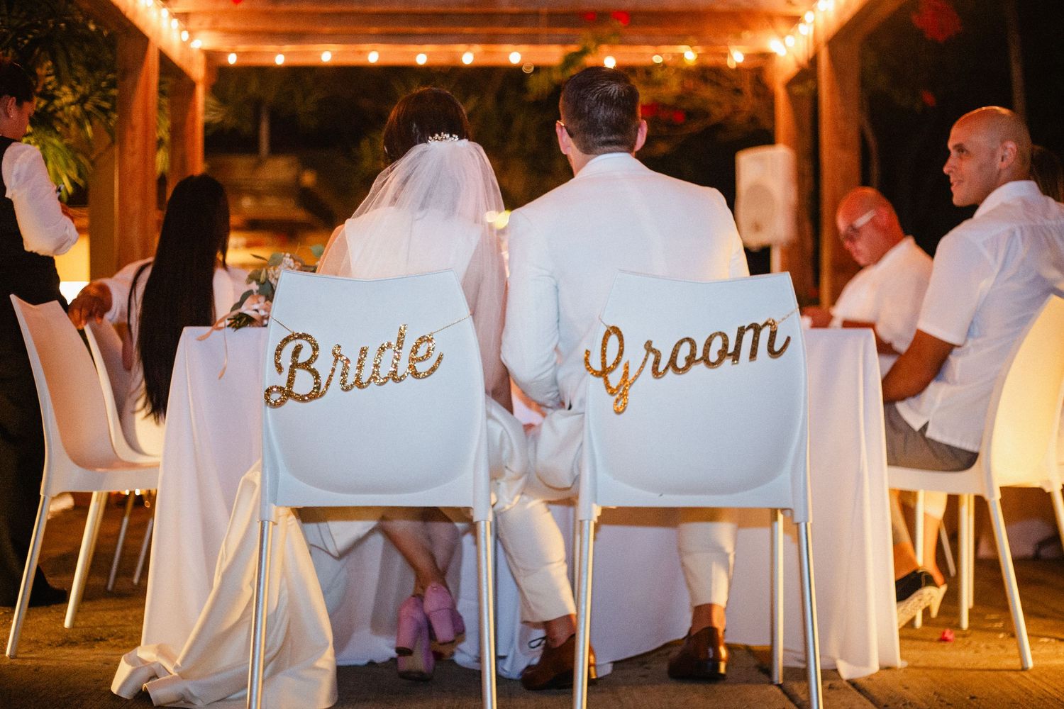 Bride and Groom sit at wedding reception table with decorative chair signs under warm string lights.