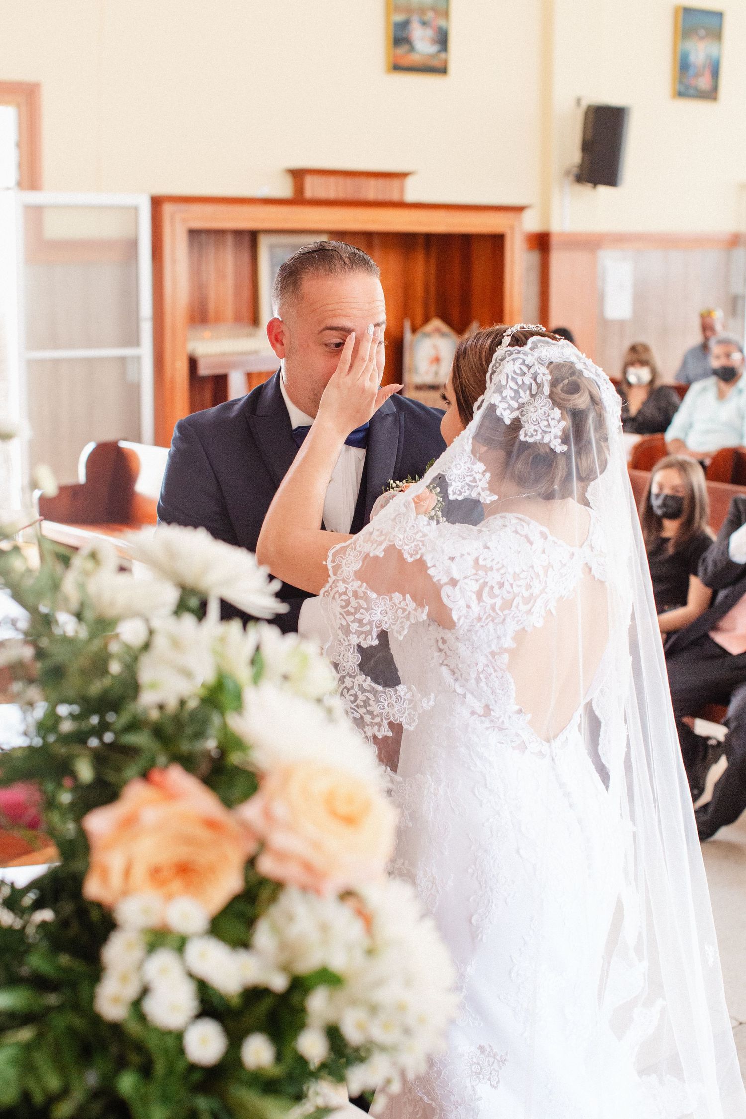 Emotional groom wiping tears as bride in lace wedding dress and veil stands before him during church ceremony.