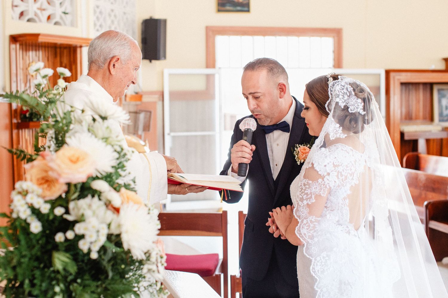 Groom reads wedding vows at church altar beside bride in lace veil, officiant holds open book amid floral arrangements.