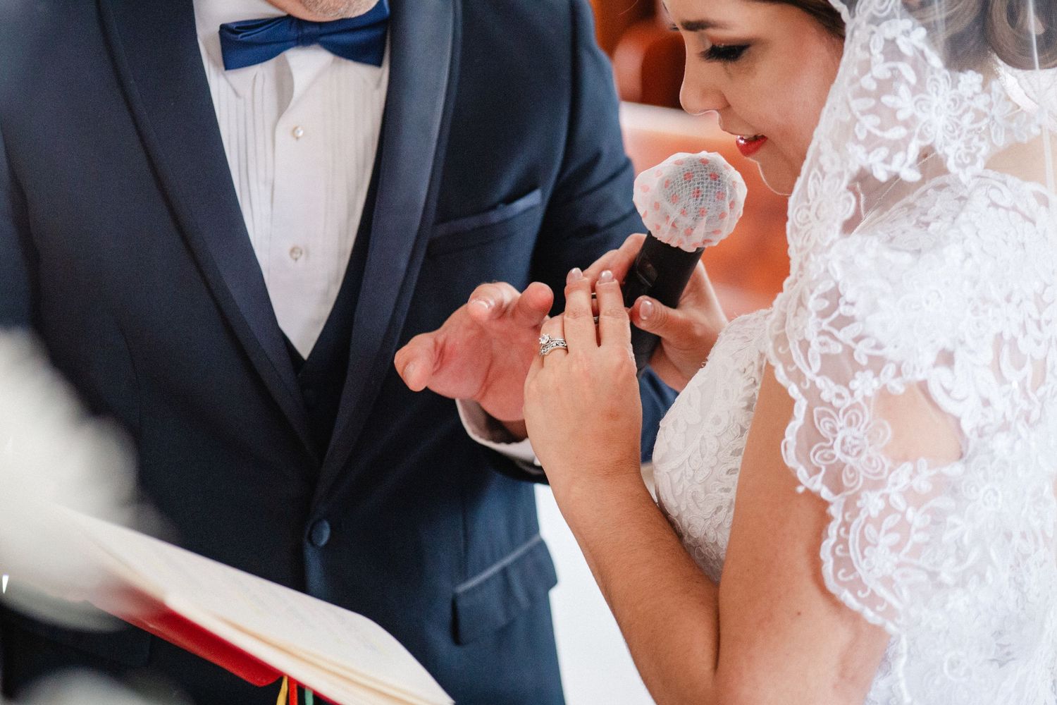 Bride placing a wedding ring on groom's finger during ceremony, wearing lace veil and white gown.