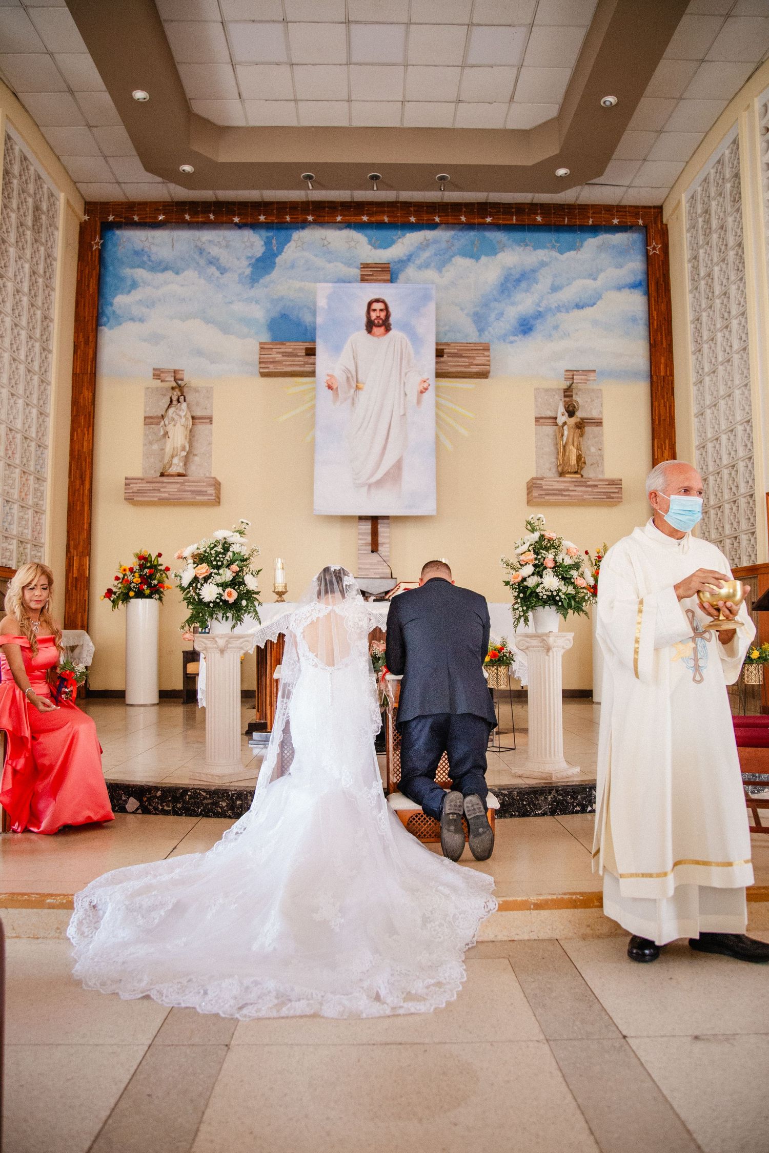 Bride and groom kneeling at church altar during wedding ceremony with priest and Jesus painting backdrop.