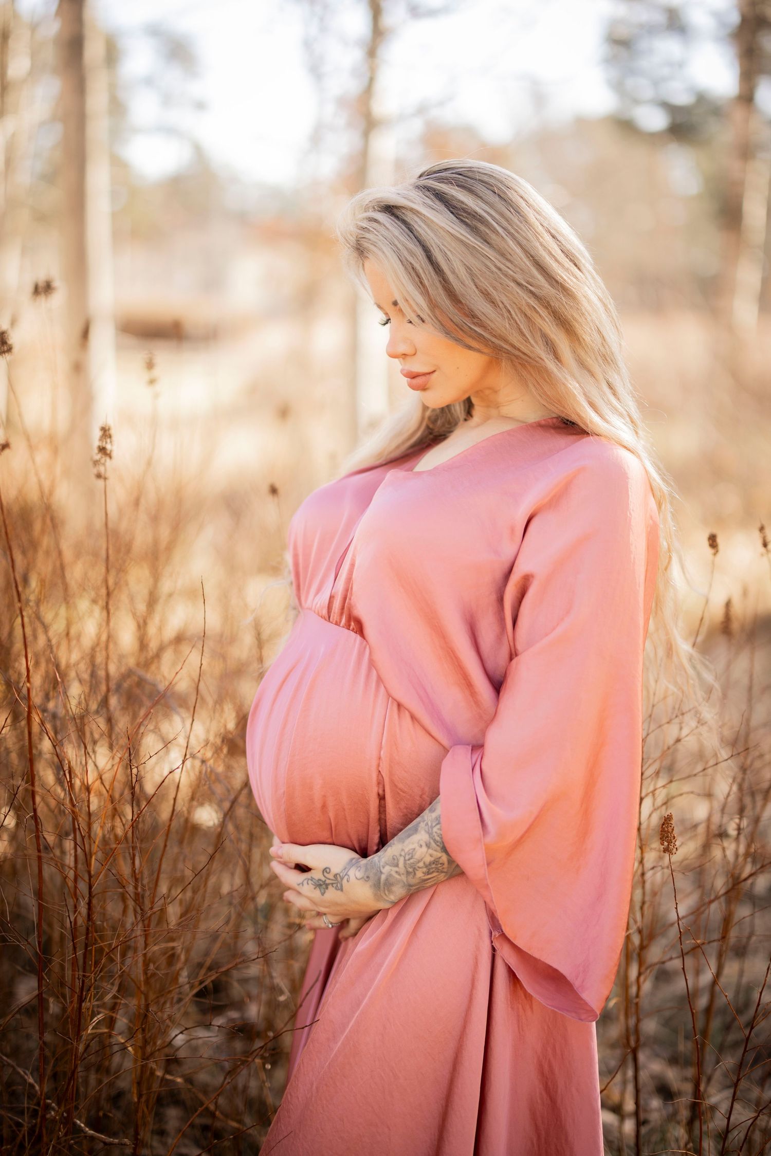 An expectant mother in a flowing rose-colored dress cradles her baby bump while standing in a sunlit field at golden hour.