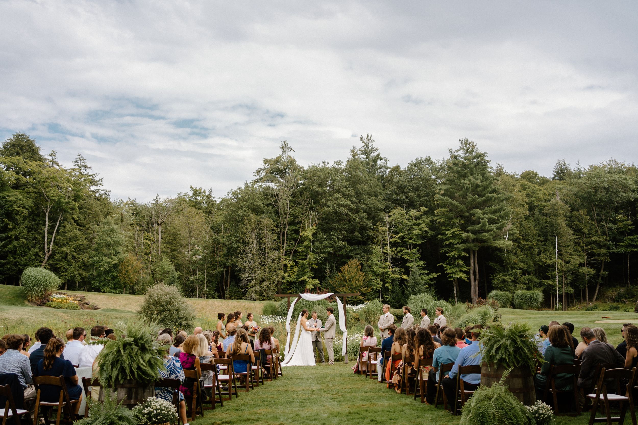 Outdoor wedding ceremony on lush green lawn surrounded by tall trees with couple at floral arch altar.