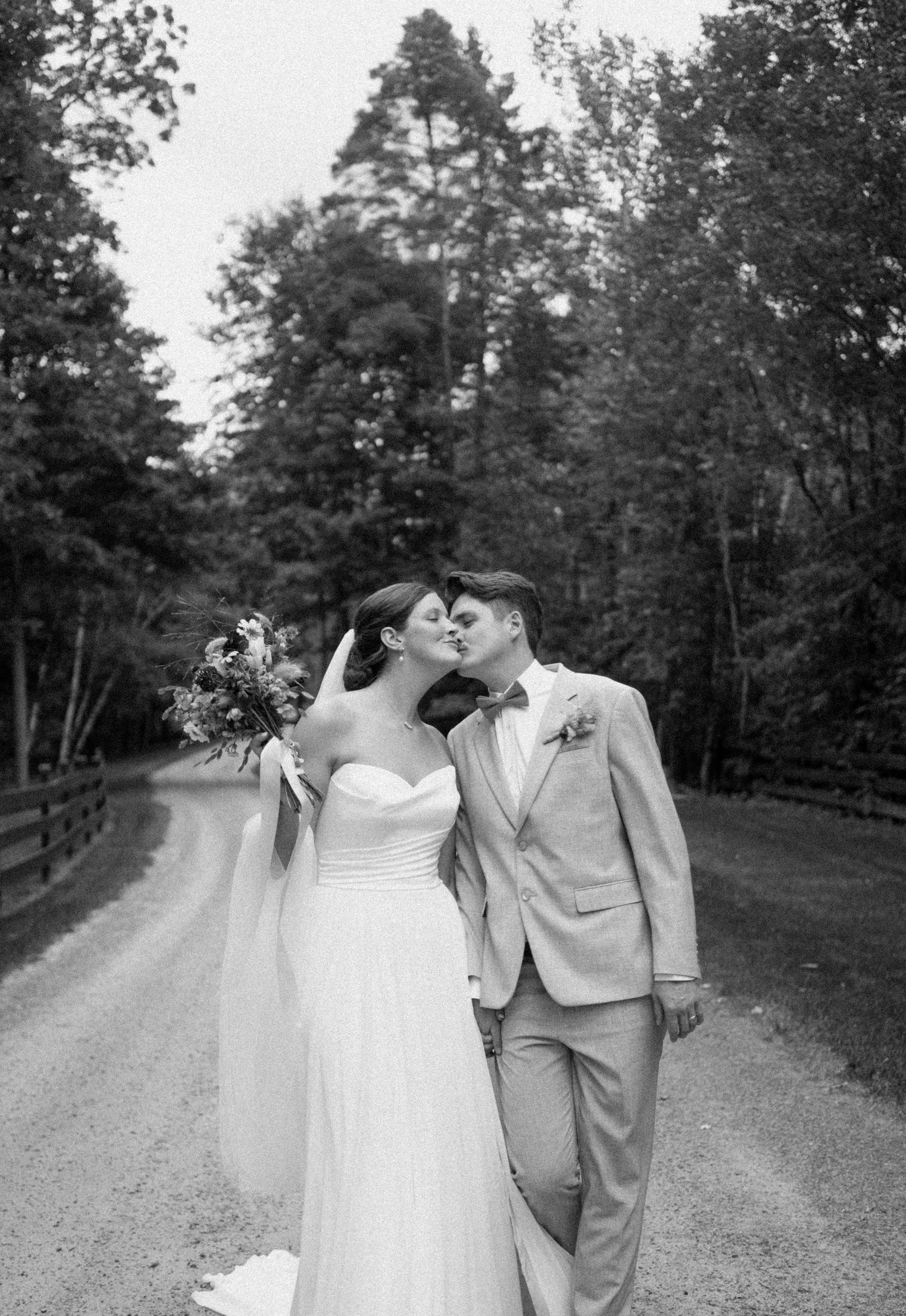 Black and white photo of a bride and groom kissing on a tree-lined path, bride holding bouquet in wedding dress.