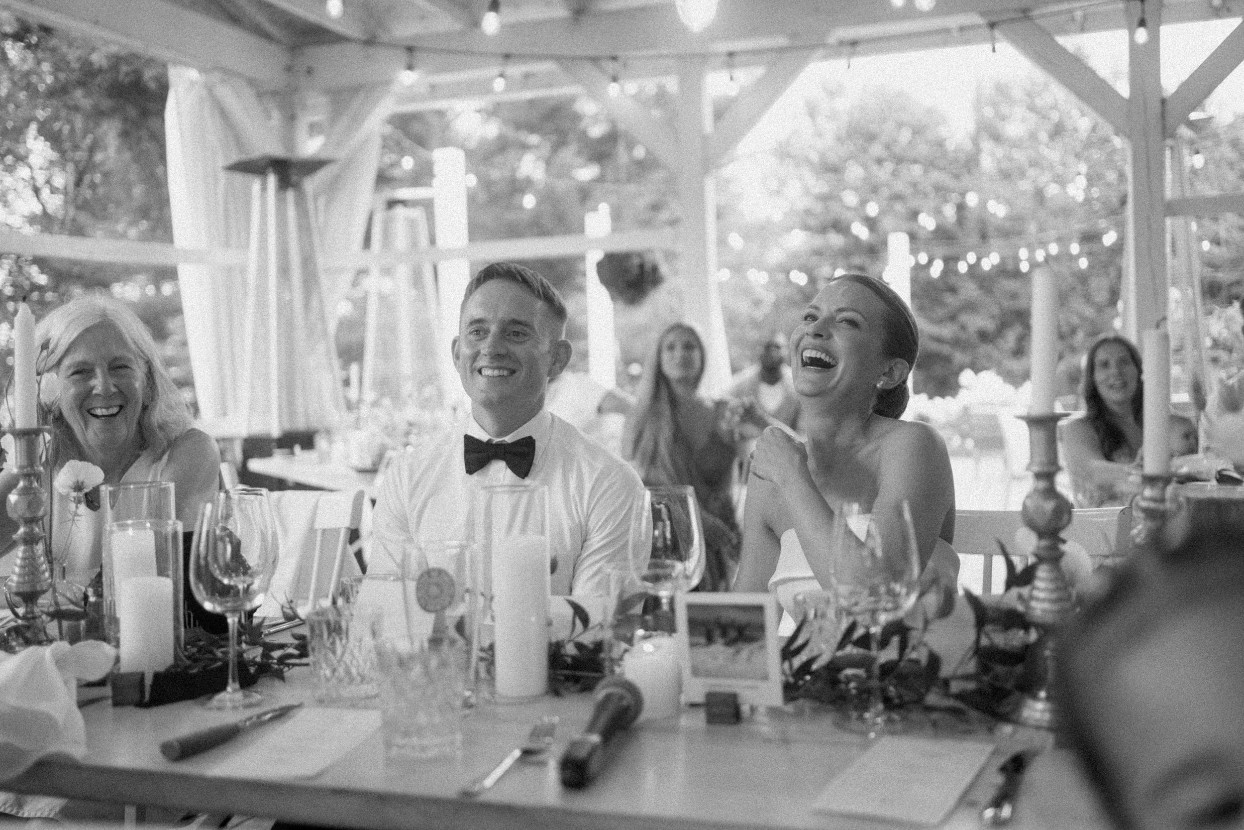 Black and white photo of wedding guests laughing joyfully at a reception table under an outdoor pavilion.