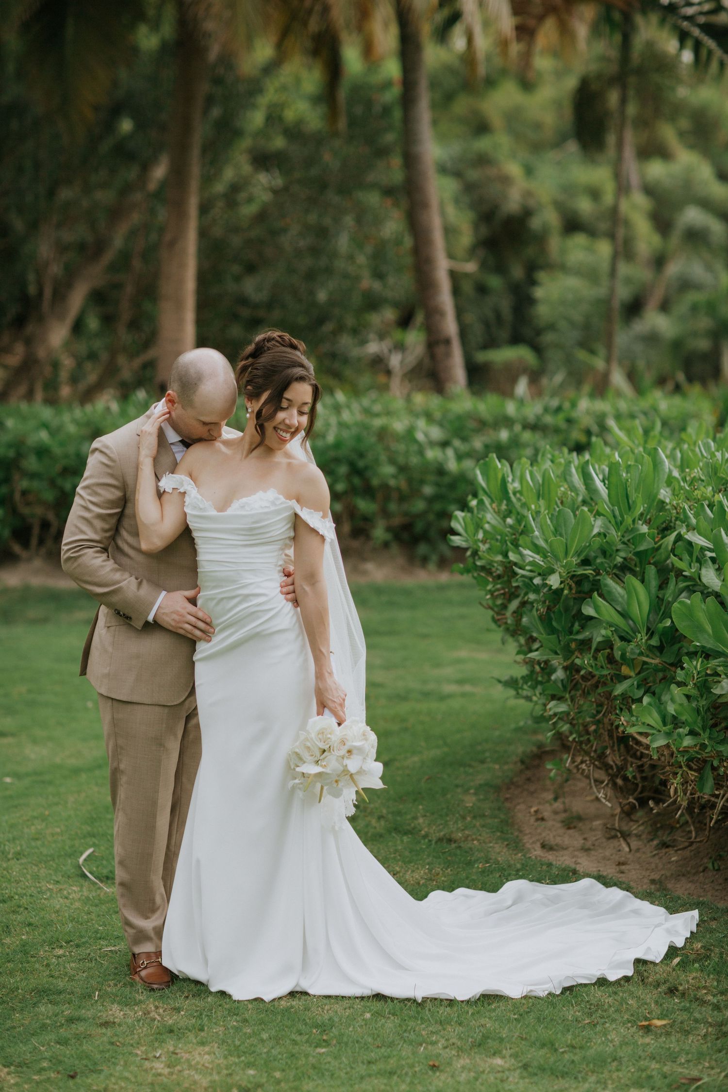 A couple shares an intimate moment in their wedding attire on a lush green lawn surrounded by trees.