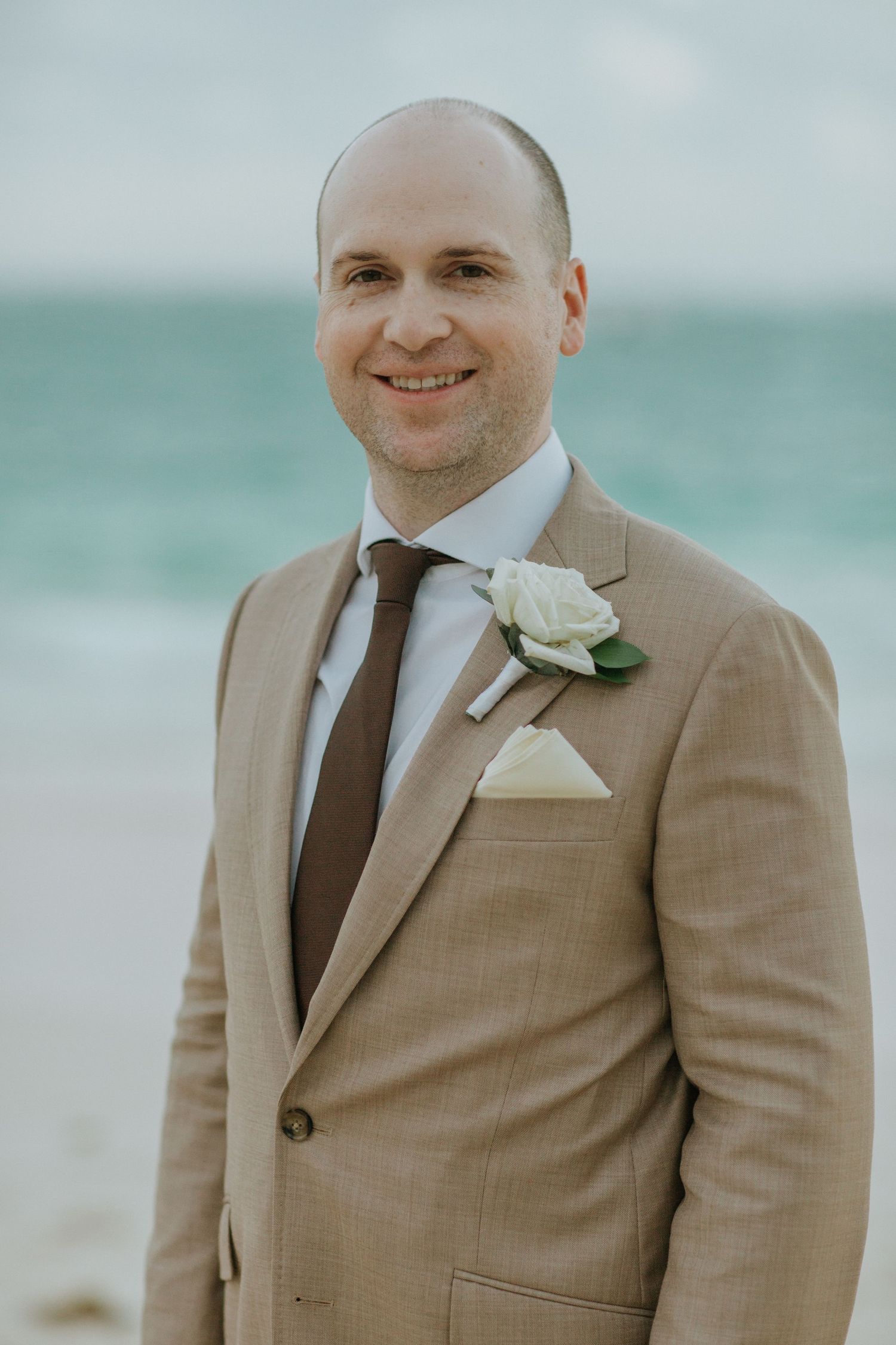 A groom in a tan suit with a brown tie and white boutonniere stands on a beach with turquoise water in the background.