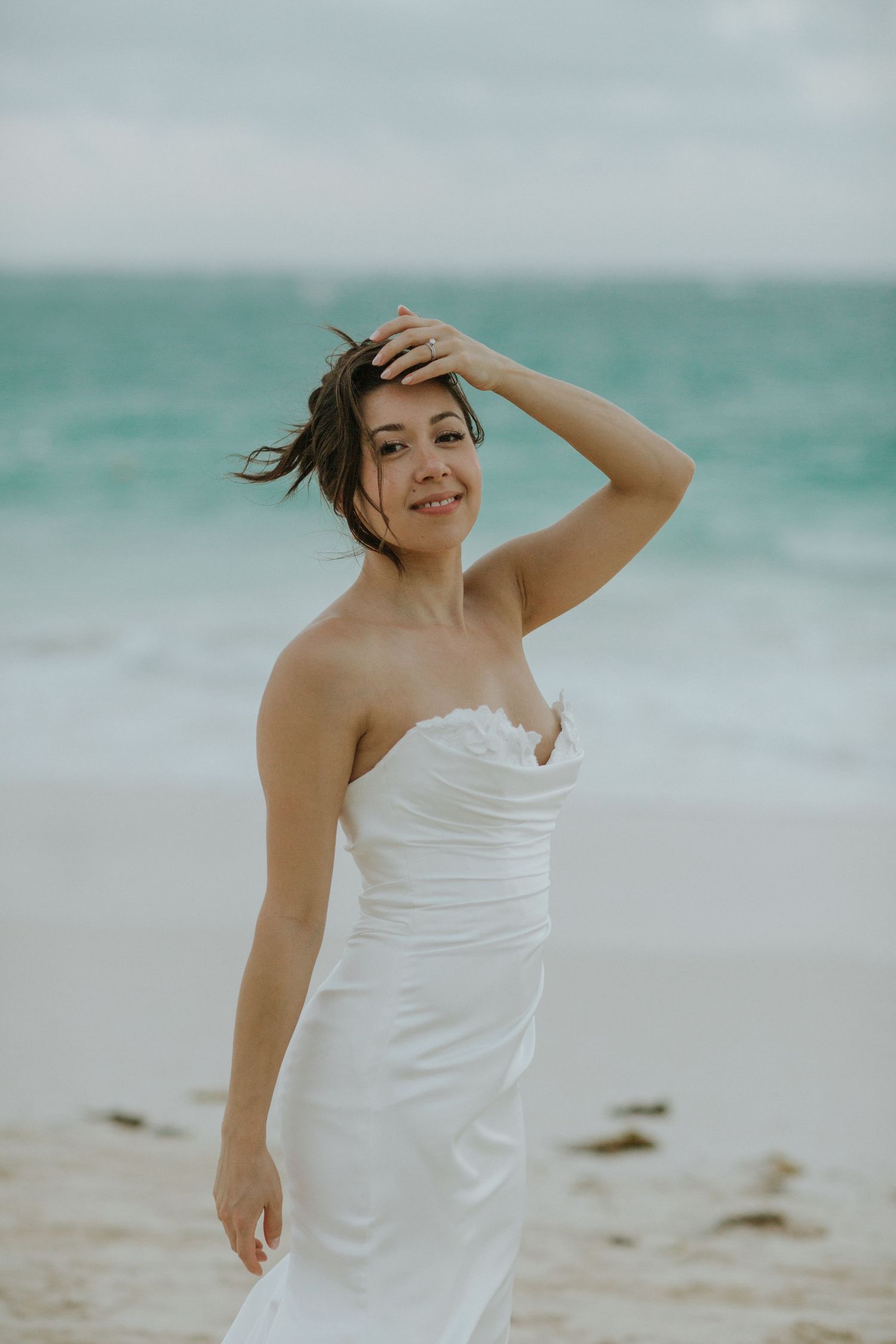 A person in a white strapless dress poses playfully on a beach with turquoise ocean waves in the background.