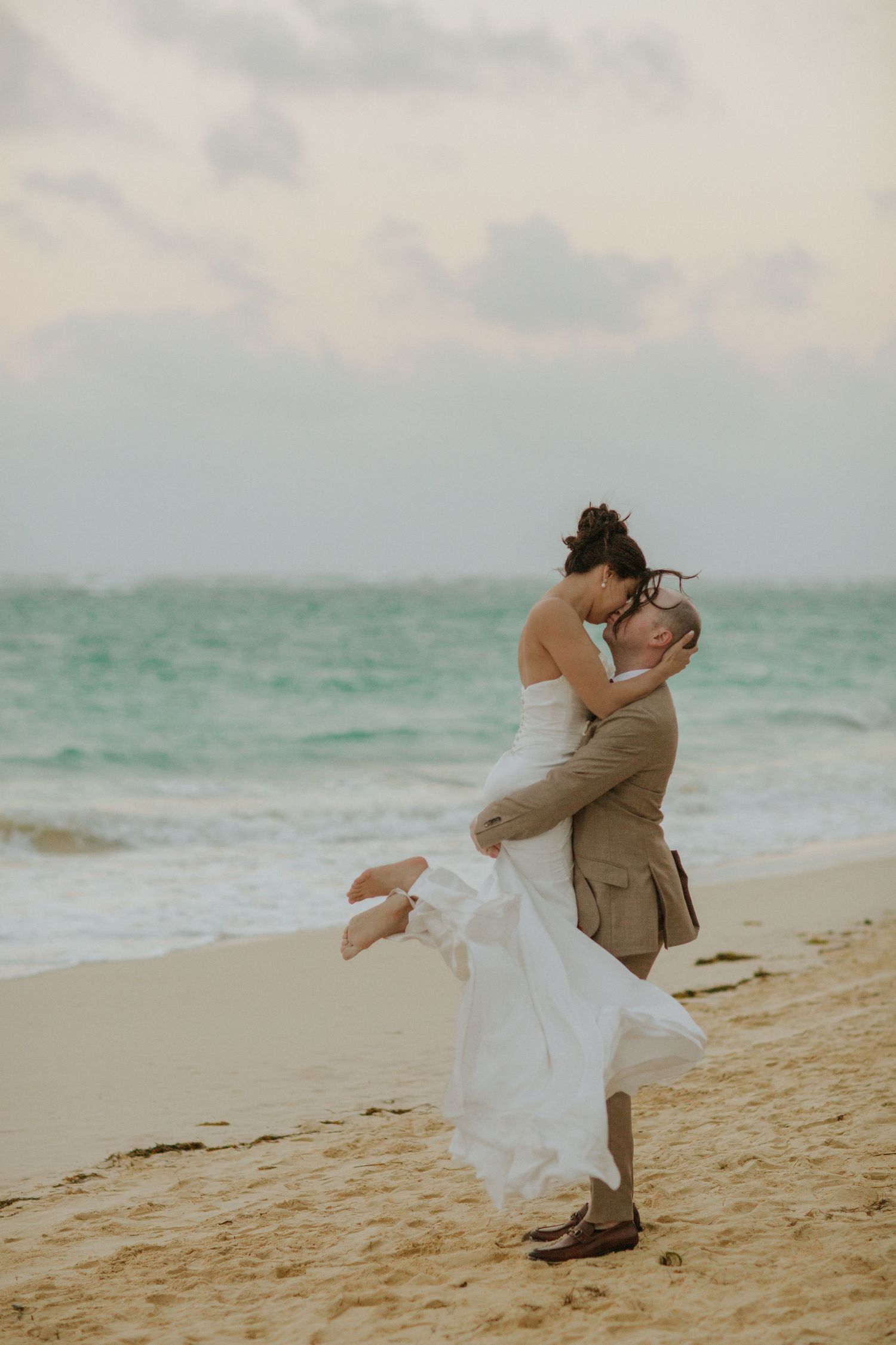 A romantic couple embraces and kisses on a sandy beach with turquoise ocean waters in the background.