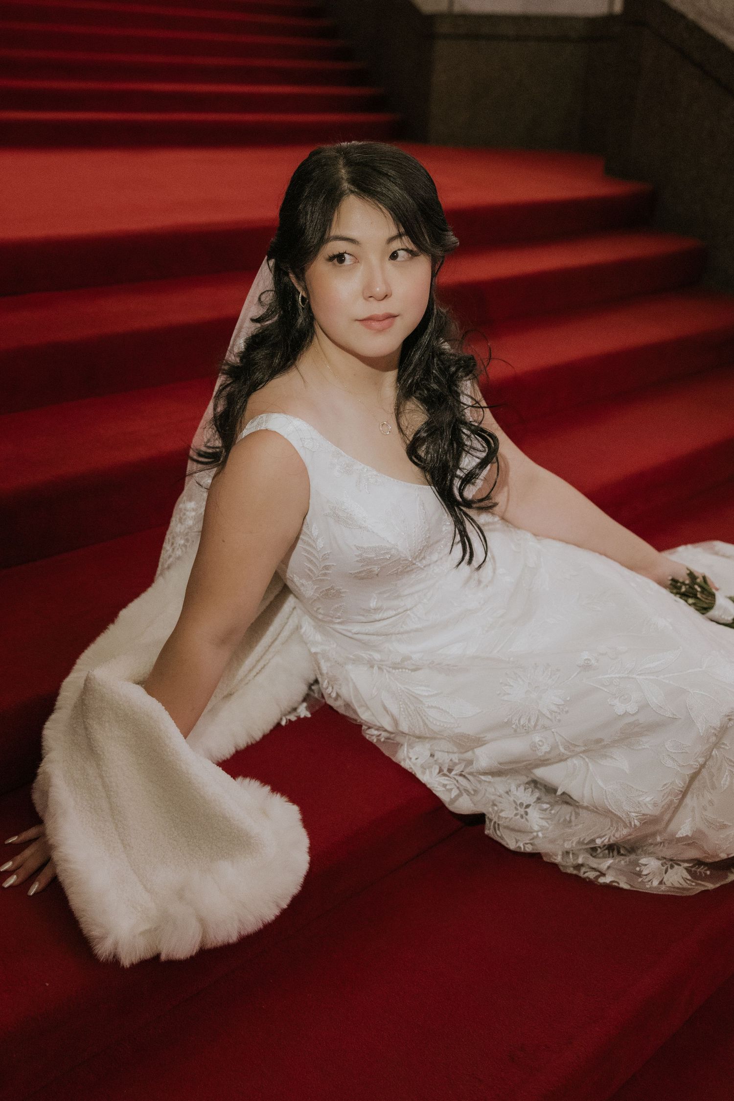 A person in a white lace gown poses elegantly on red carpeted stairs in a romantic bridal photoshoot.
