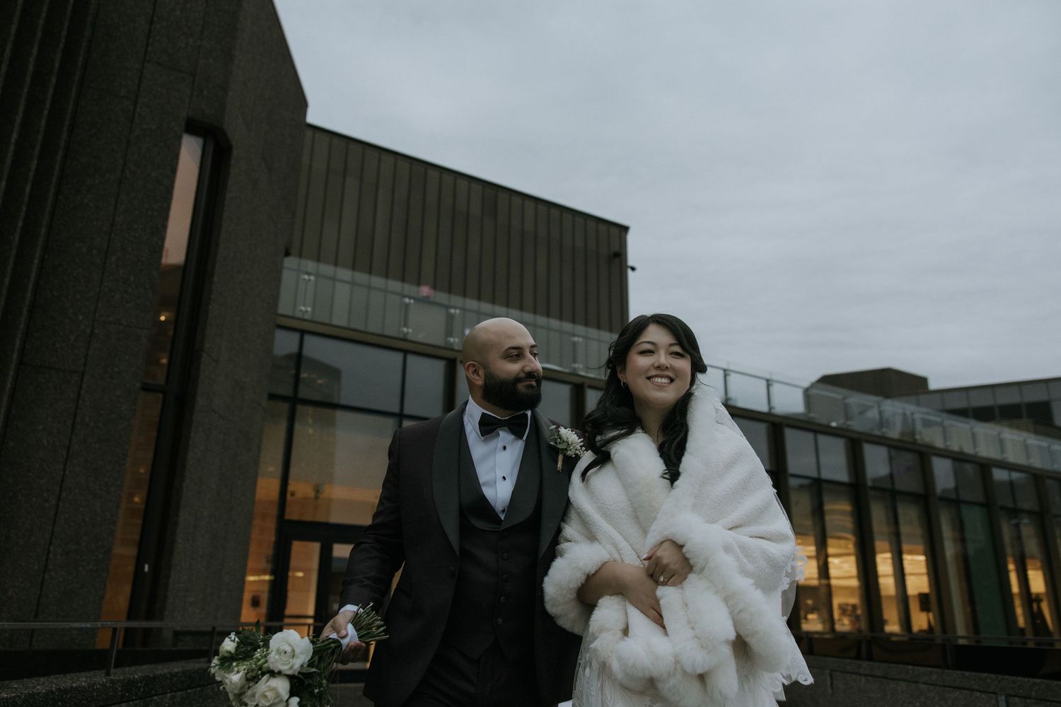 A couple poses for wedding photos outside a modern building on an overcast day.