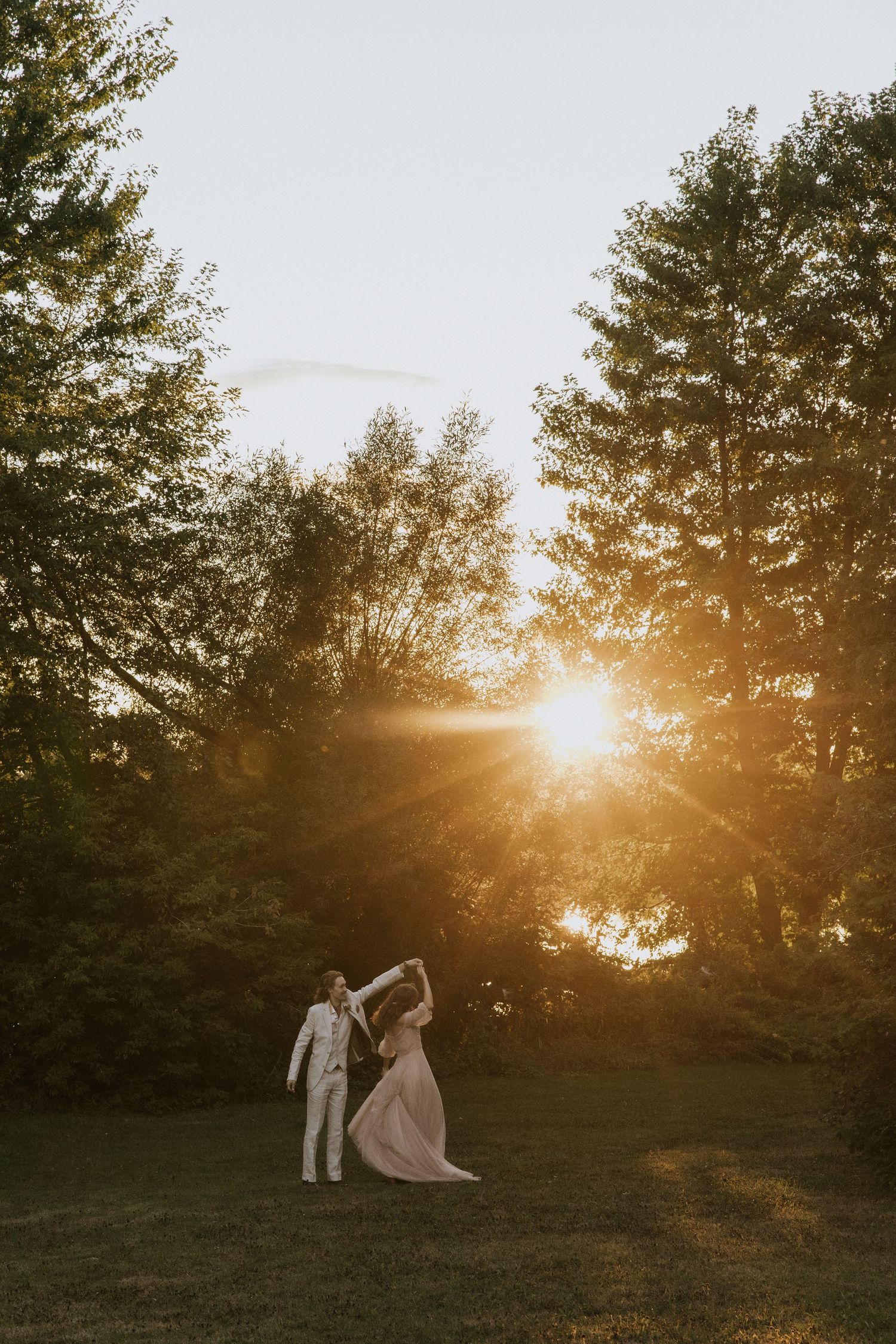 A romantic sunset silhouette of a couple dancing on a grassy lawn surrounded by tall pine trees.