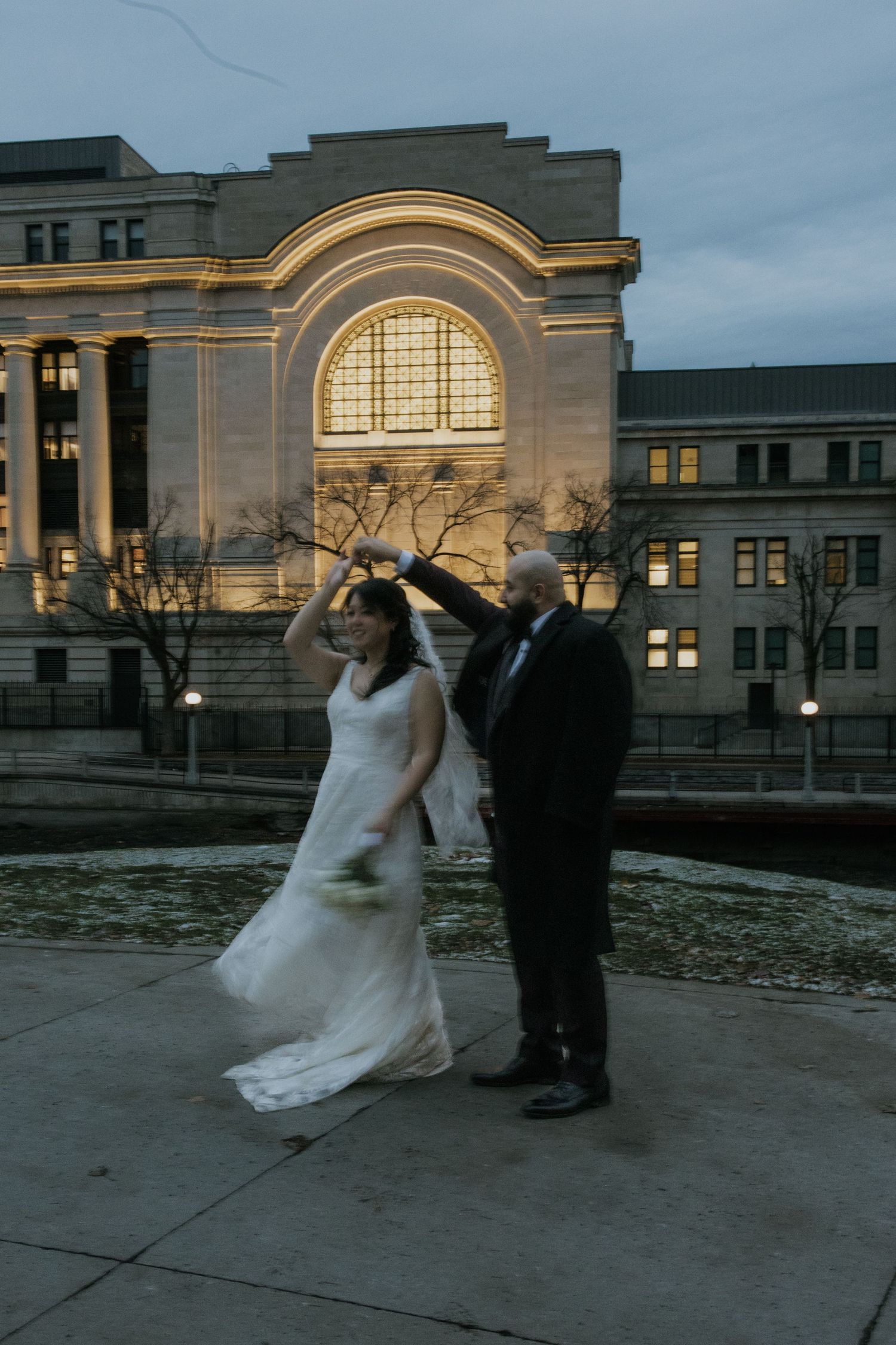 A couple in wedding attire dances outside a historic building at dusk with illuminated arched windows in the background.