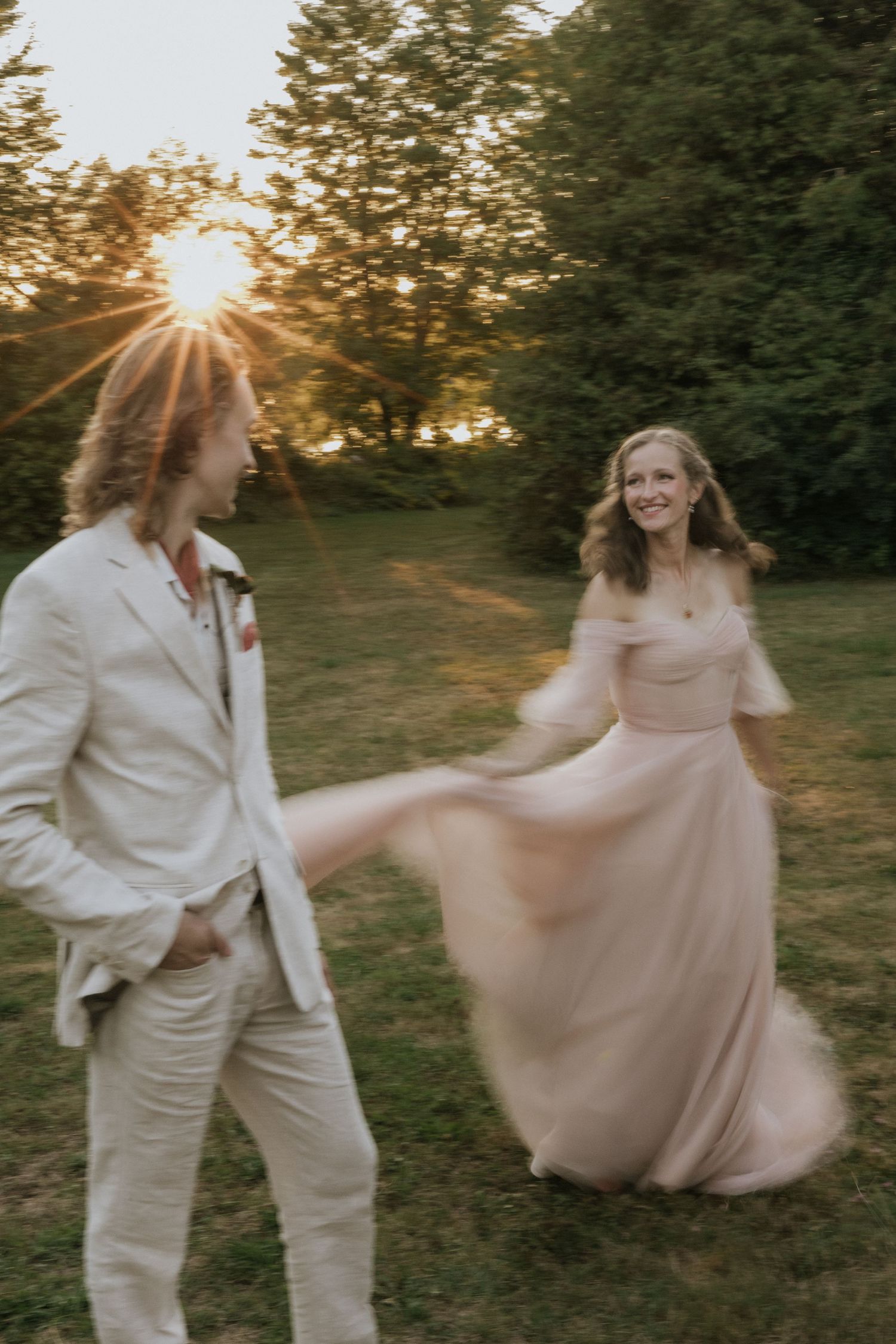 A couple dances together in a grassy field at sunset, with one wearing white and the other in a flowing blush dress.