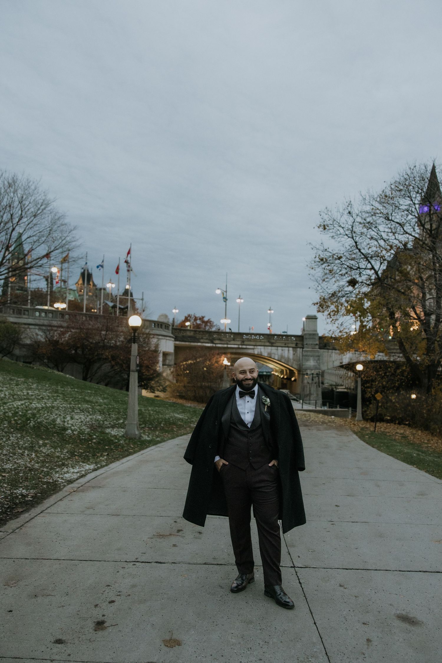 A person in a dark coat walks along a concrete path on an overcast autumn day with bare trees and flags in the background.