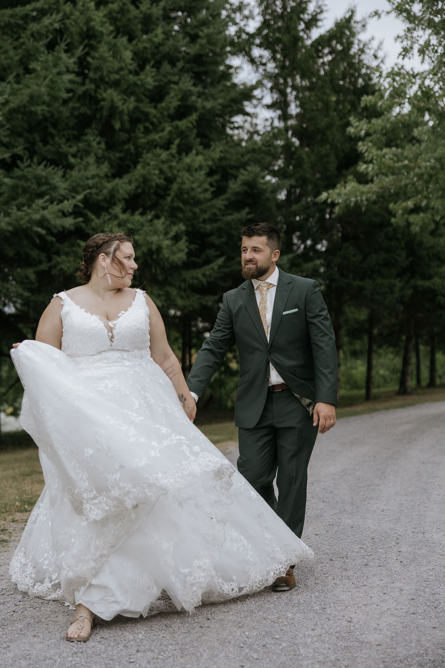 A romantic wedding couple walks and plays on a gravel path surrounded by green trees in a park setting.