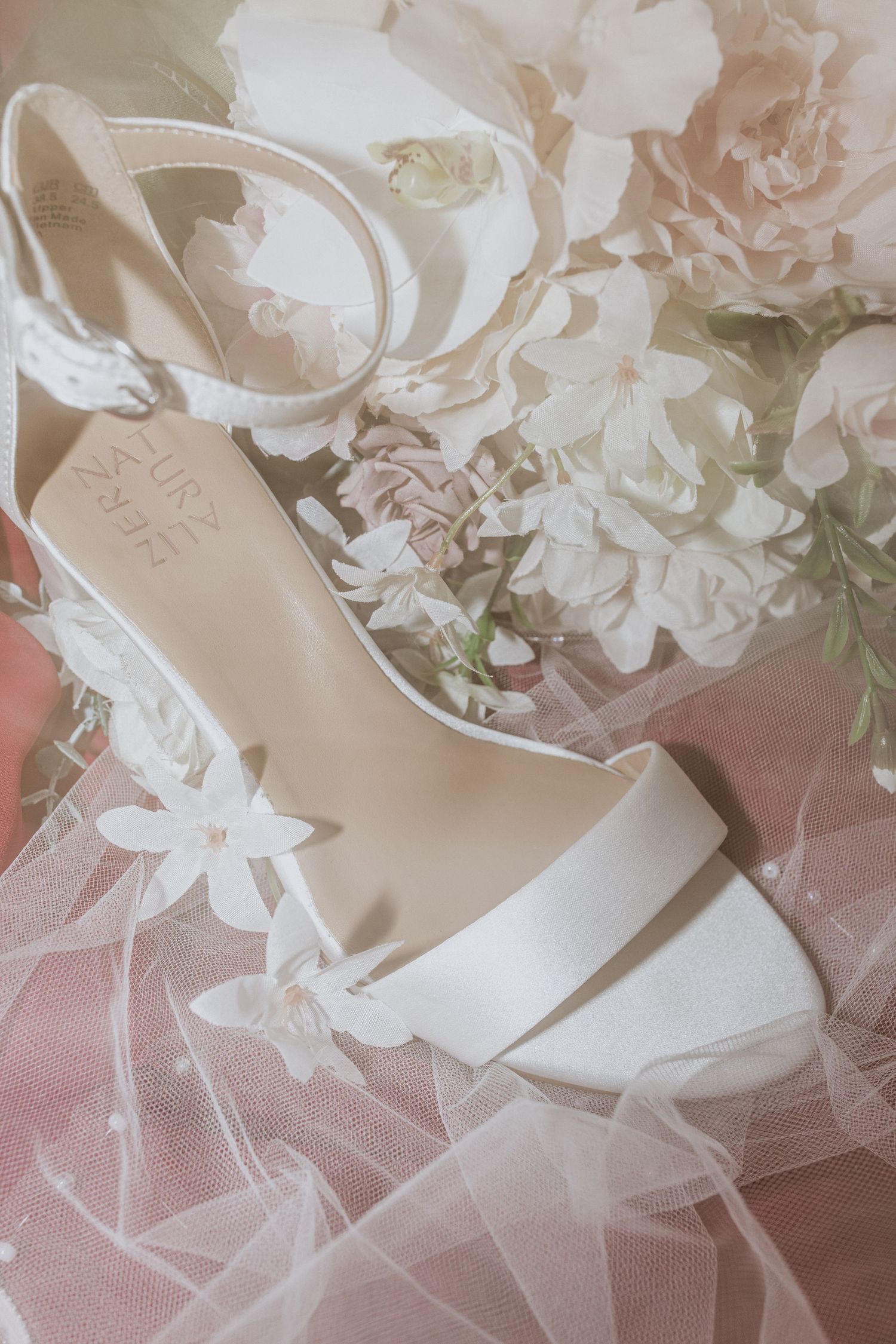 Elegant white bridal sandal with ankle strap rests on soft tulle fabric and delicate floral details.