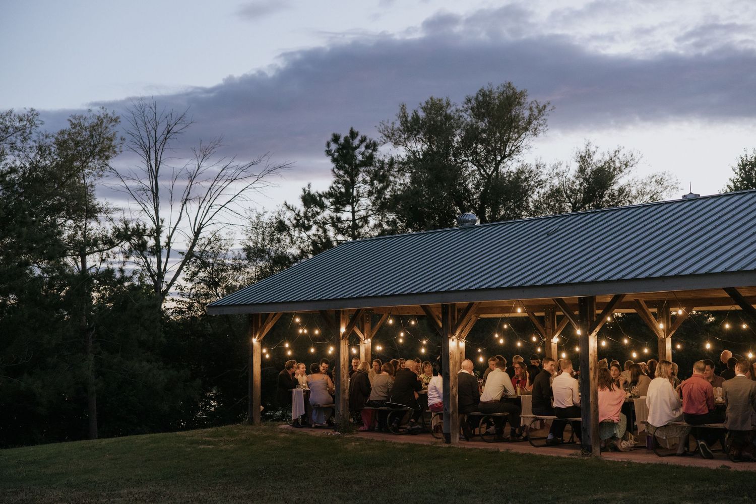 Outdoor wedding reception under a covered pavilion at sunset with string lights and decorative floral arrangements.