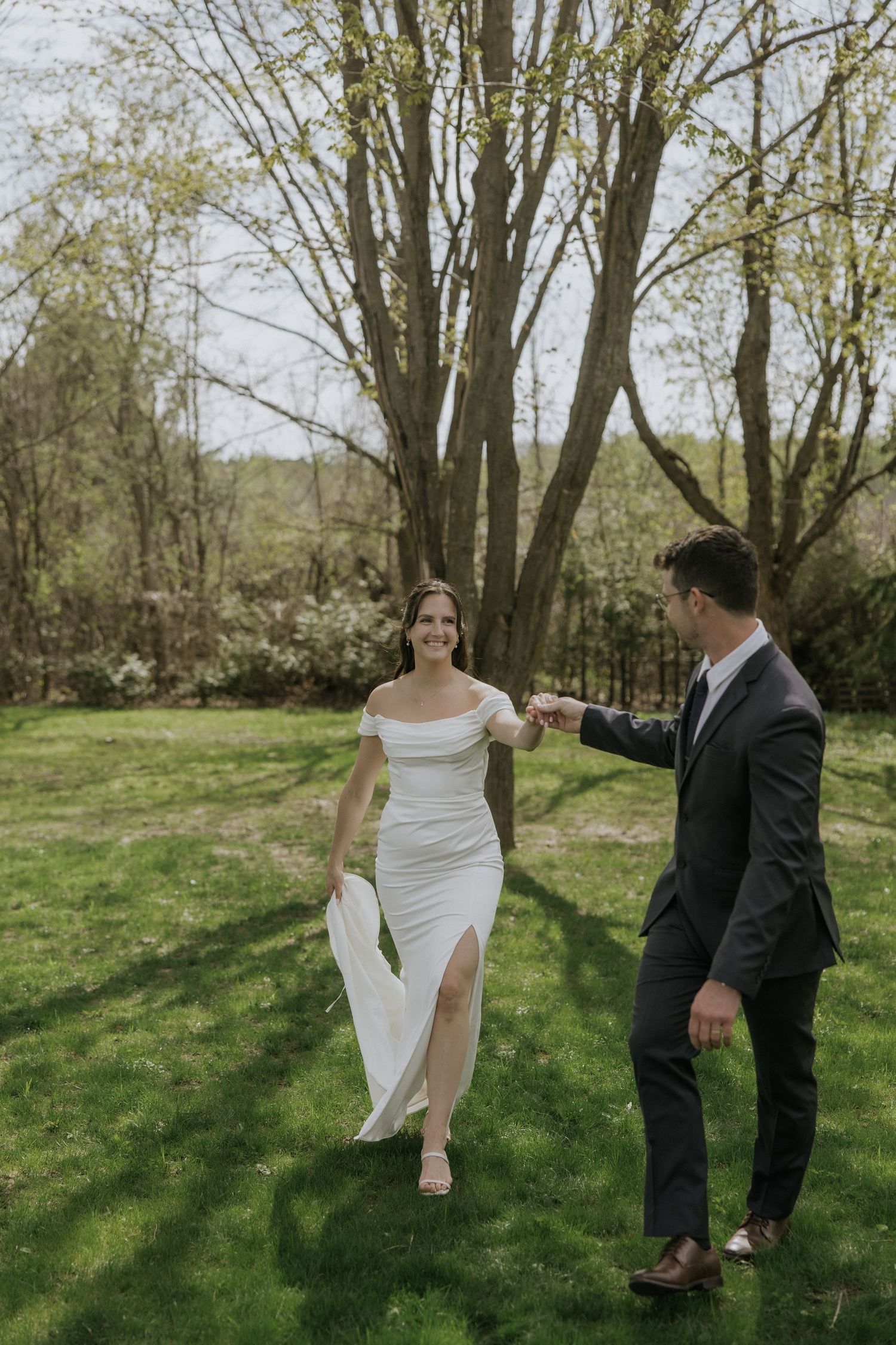 A couple in wedding attire playfully dances together in a sunlit wooded area on their special day.