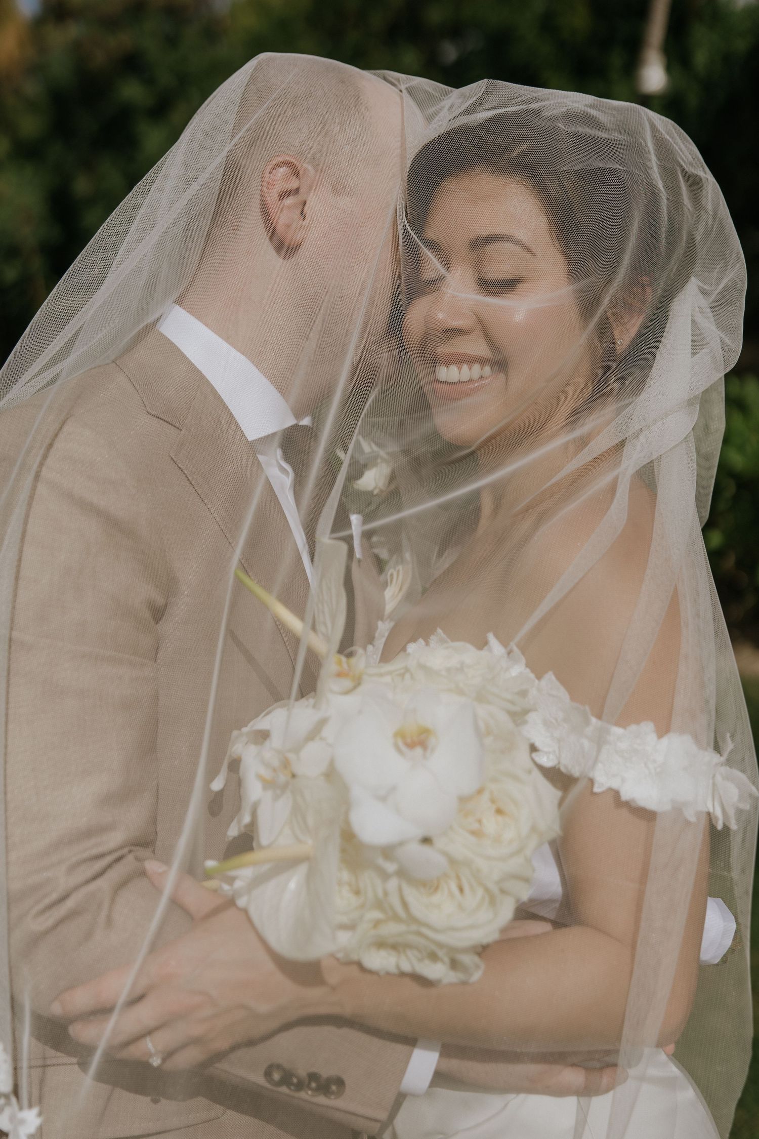 A romantic wedding couple embraces under a delicate white veil in an outdoor garden setting.