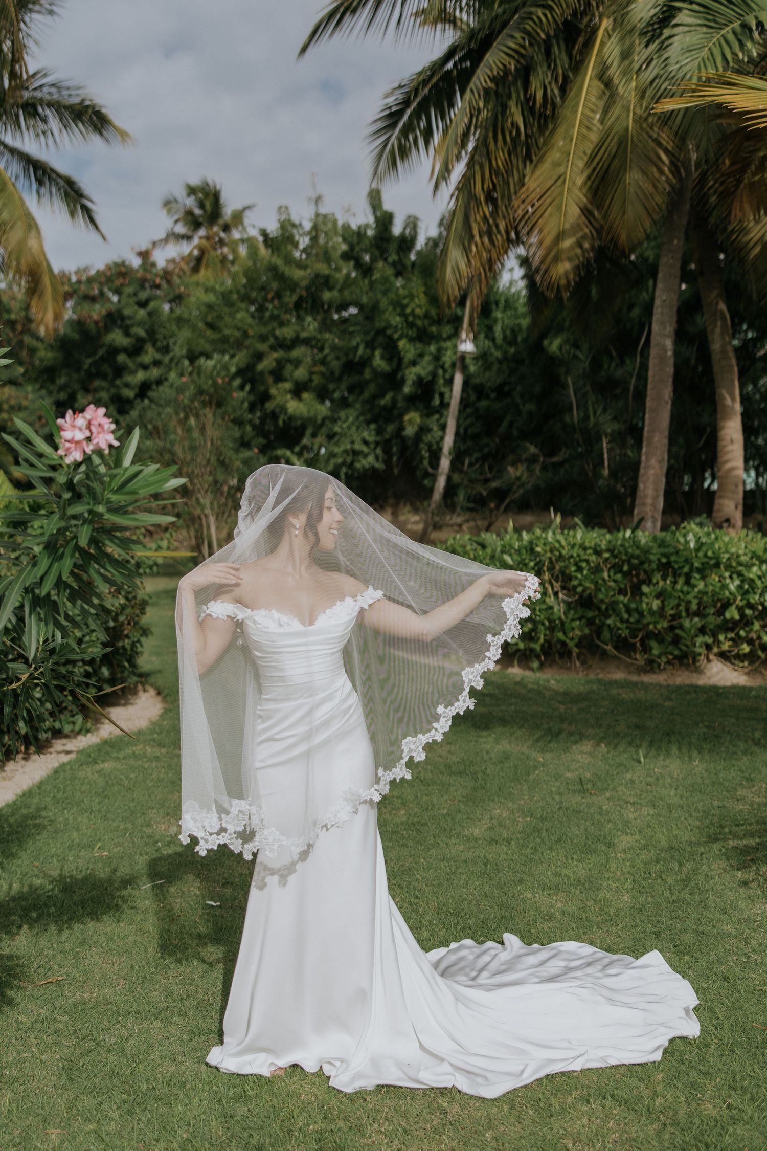 A flowing white wedding dress with lace veil billows gracefully on a green lawn with palm trees in the background.