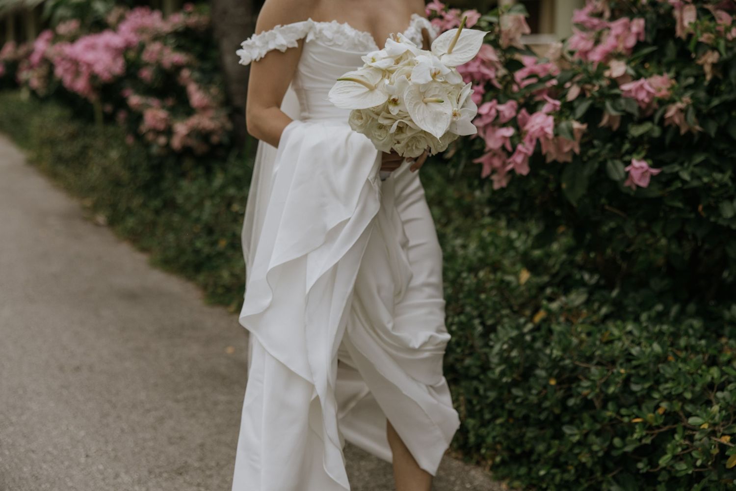 A white wedding dress with lace details flows elegantly against a backdrop of pink flowering bushes.