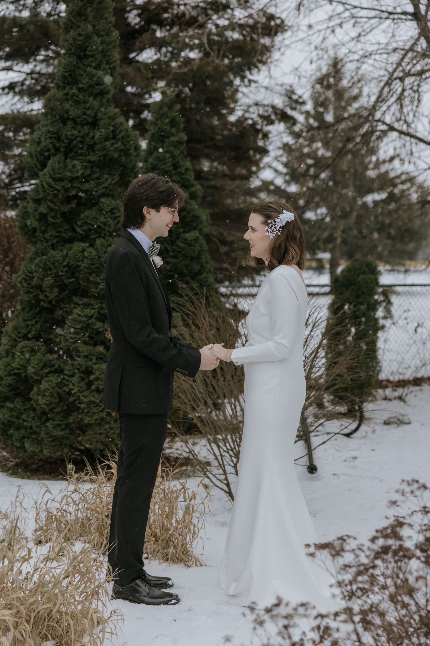A couple shares an intimate winter moment among snow-covered evergreen trees and dried grasses by a lake.
