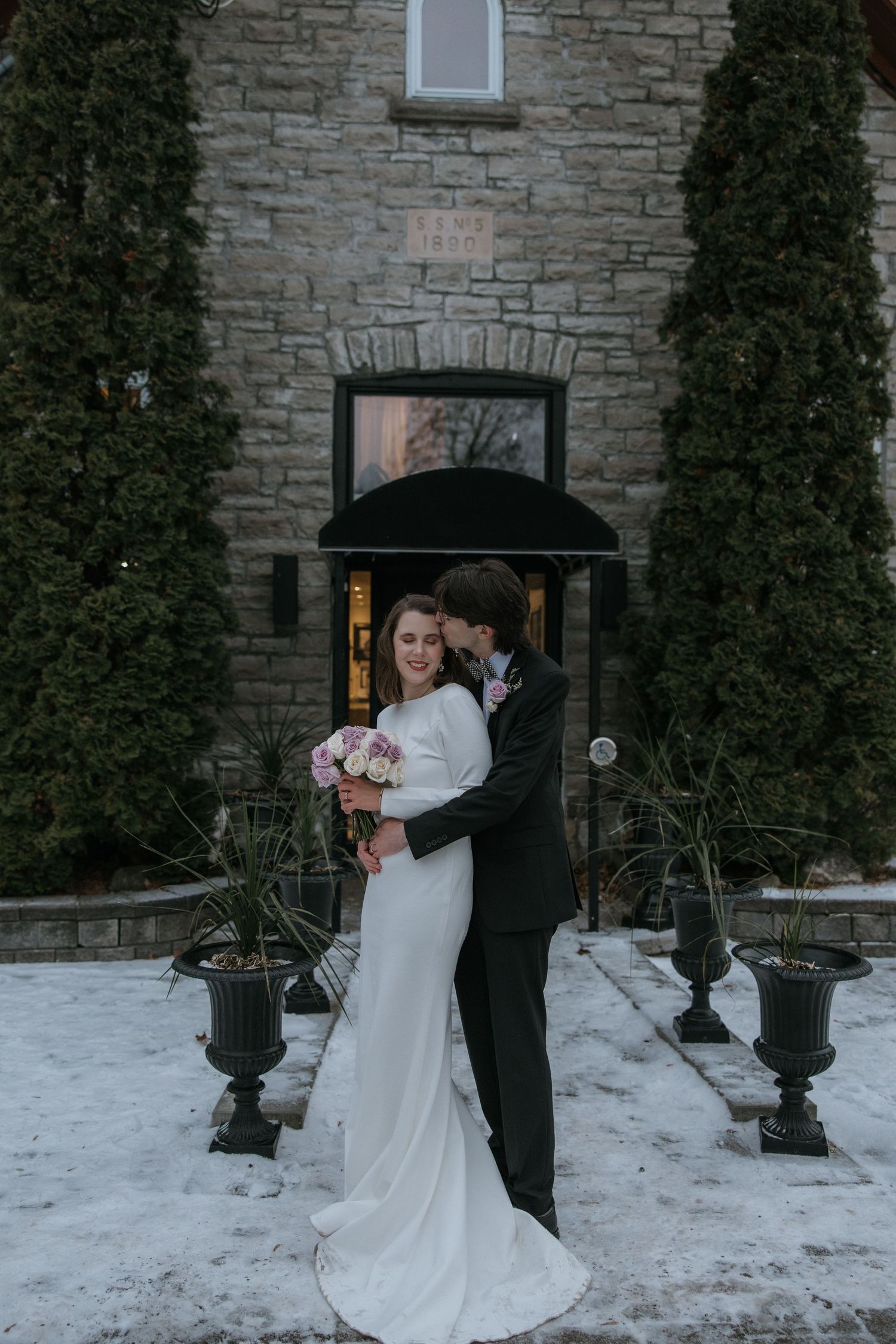 A romantic winter wedding couple embraces against a stone wall backdrop in the snow at dusk.