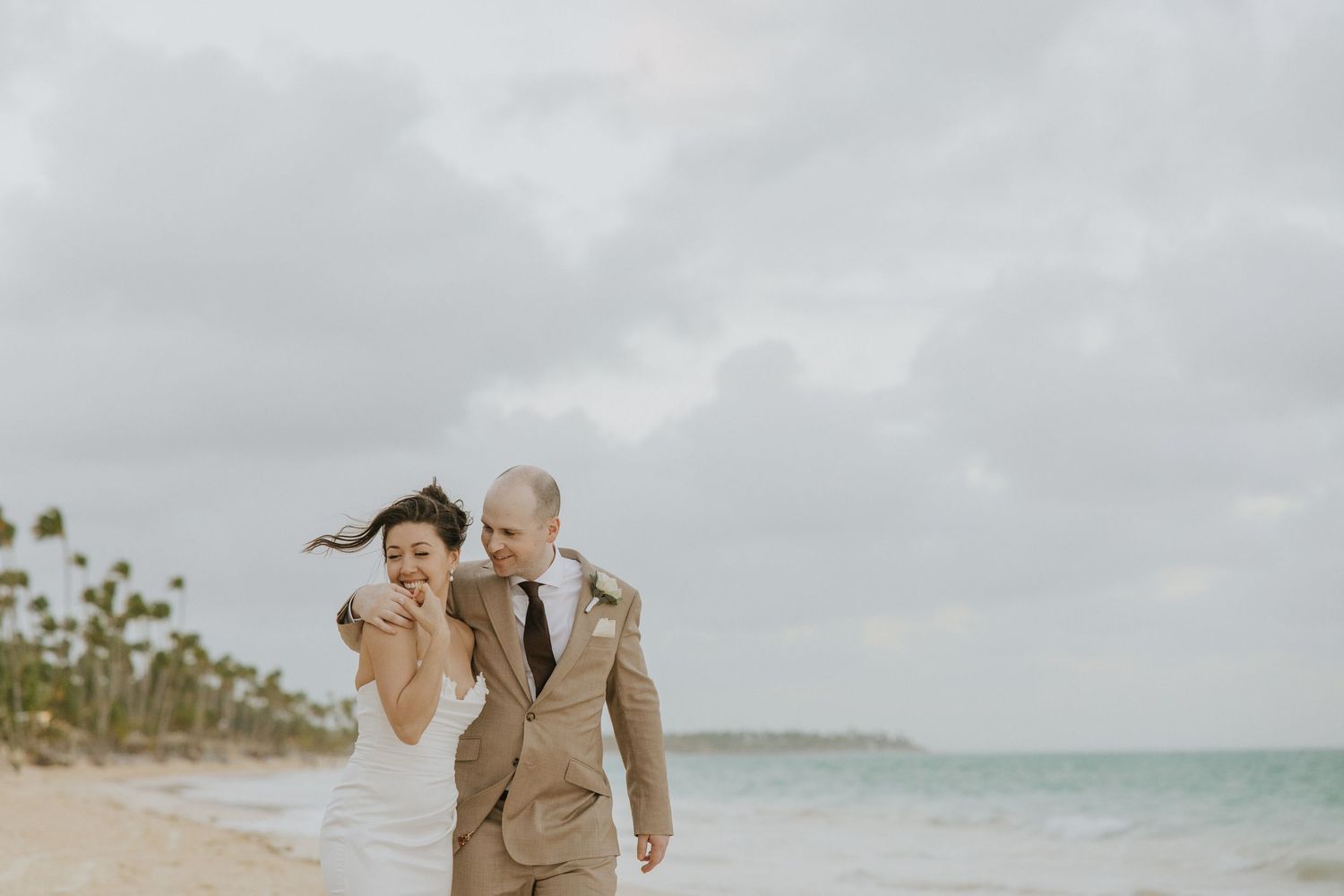 A couple in khaki outfits walks along a tropical beach with palm trees and turquoise water in the background.