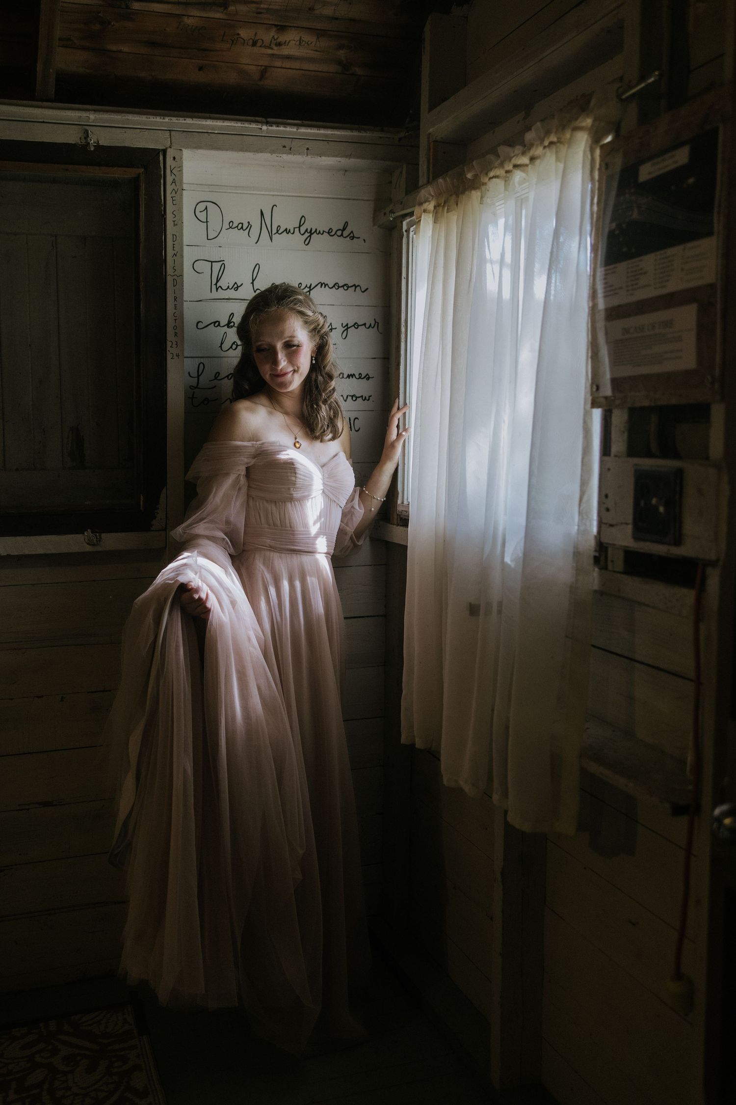 A figure in a flowing pink vintage dress stands in a dimly lit rustic cabin interior near a window.