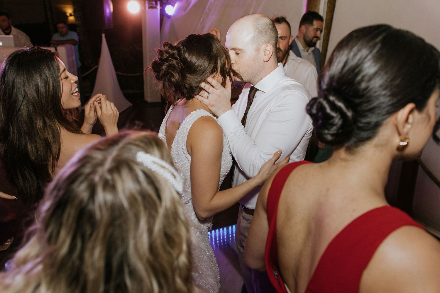 People dancing and celebrating at a dimly lit wedding reception with red and purple lighting.