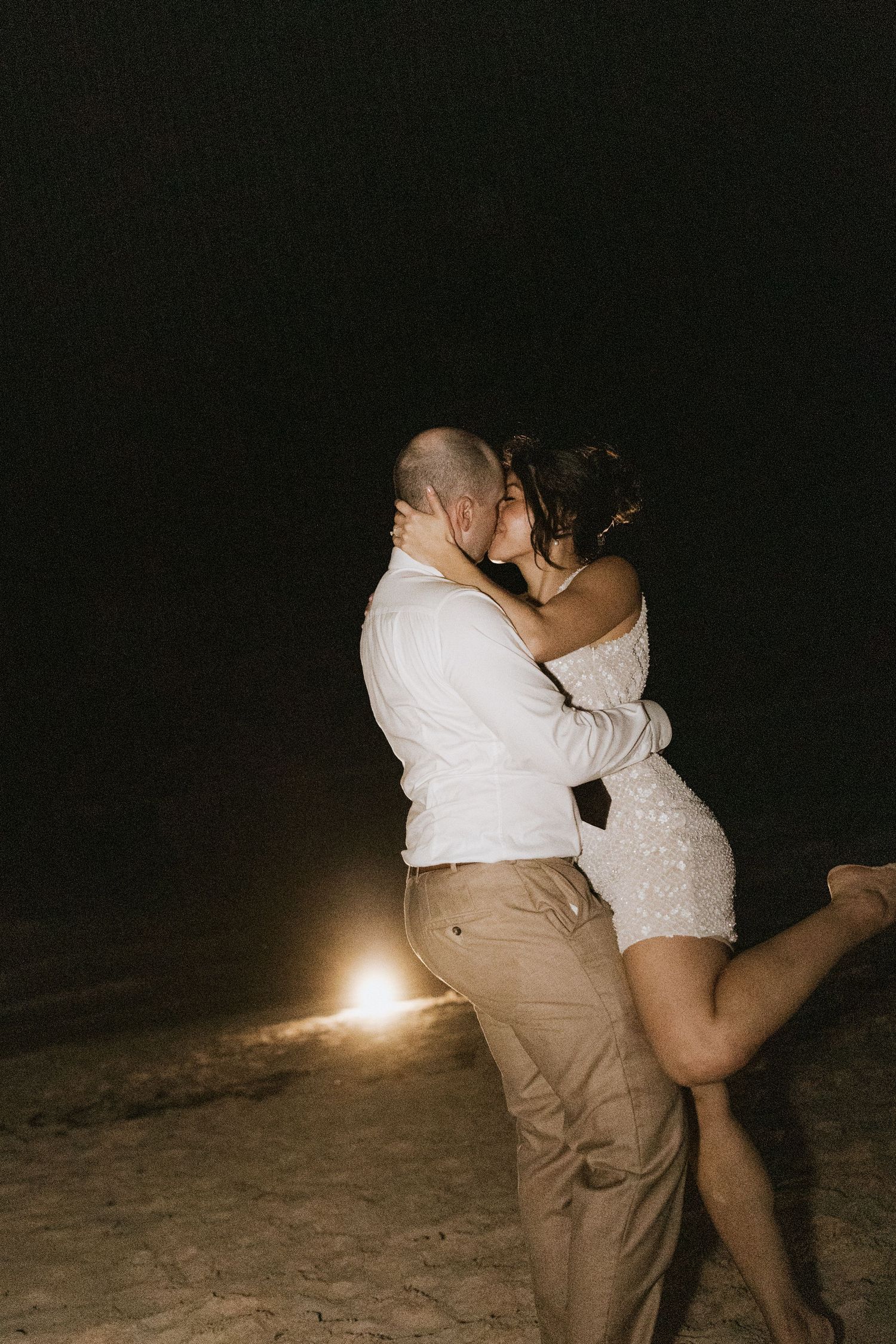 A romantic couple embraces in a white dress and khakis on a dark beach with a single light glowing in the background.