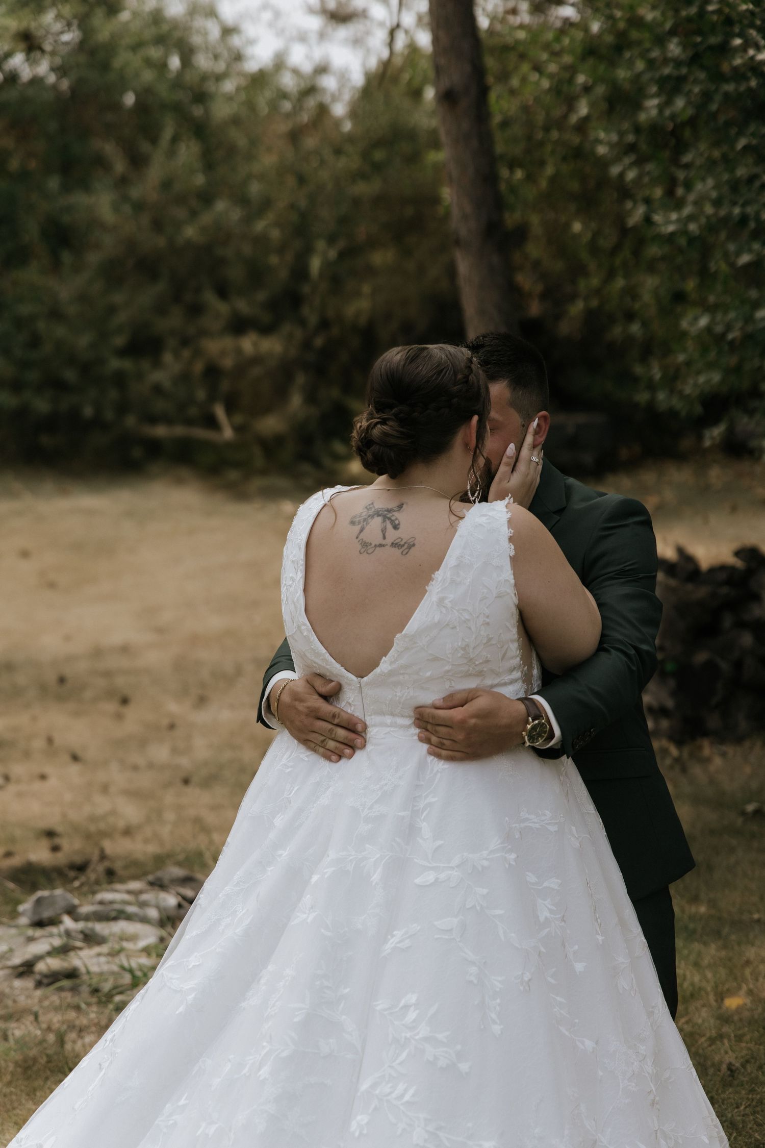 A romantic wedding moment showing the back of a bride in a white lace gown with a visible tattoo embracing her partner outdoors.