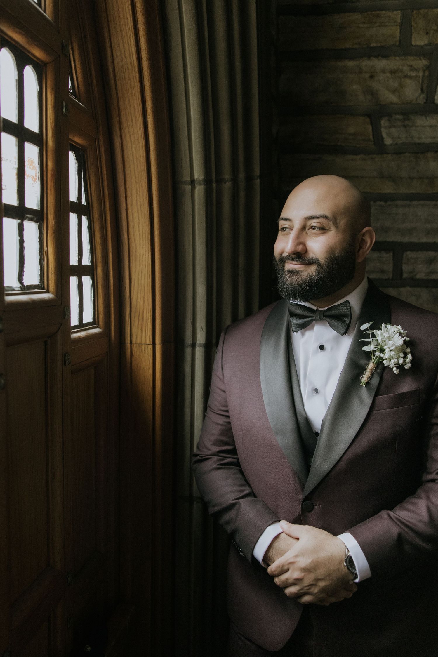 Groom in burgundy tuxedo and white boutonniere poses in moody window light for wedding portraits.