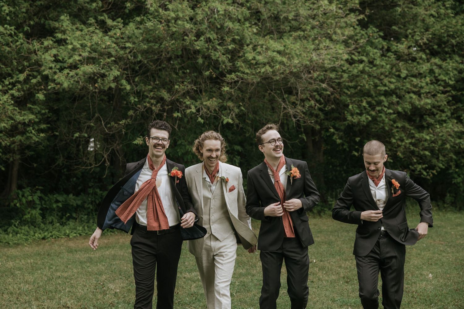 Wedding party members in black suits and orange ties walk together joyfully on a grassy path surrounded by trees.