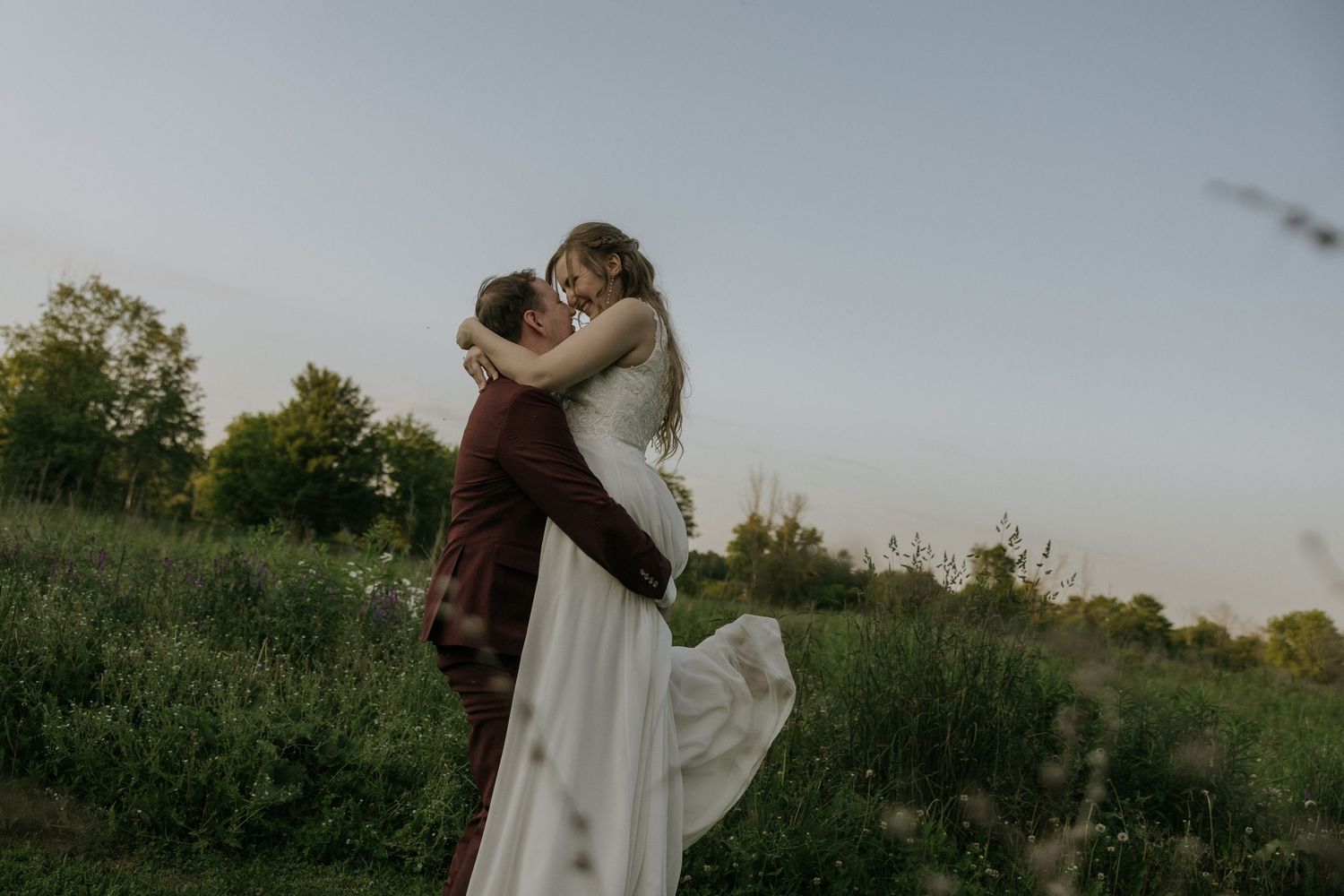 A romantic wedding couple shares an intimate moment in a rustic outdoor field at sunset.
