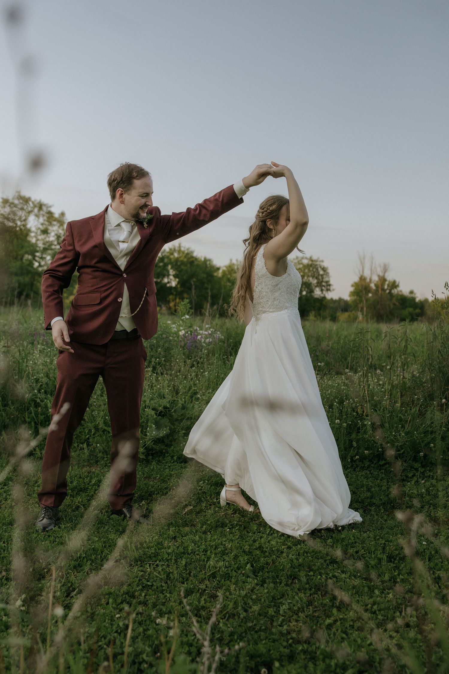 A couple dances together in a meadow at sunset, with the bride in white and groom in burgundy suit.