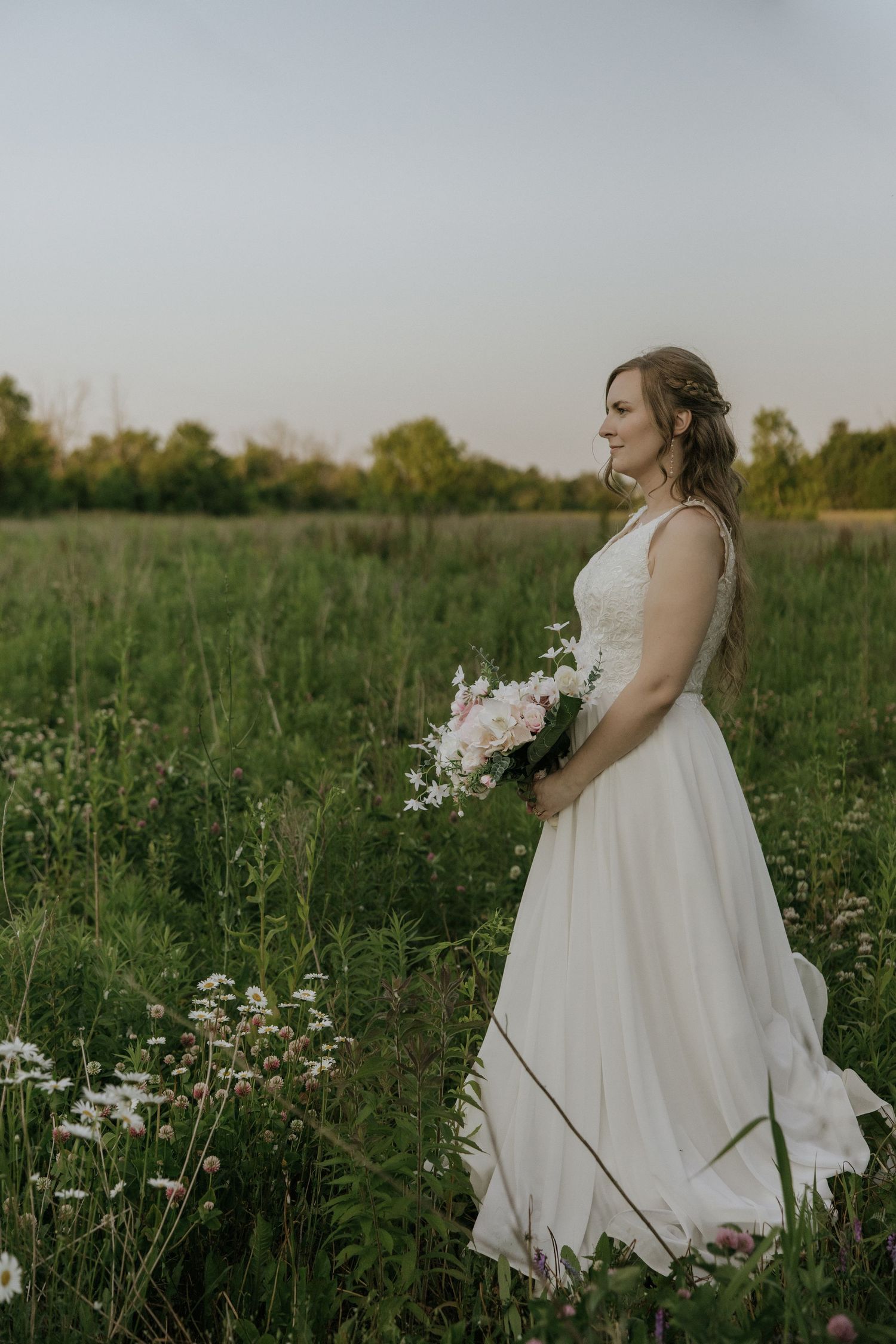 A bride in a flowing white dress holds a bouquet while standing in a wildflower meadow at dusk.