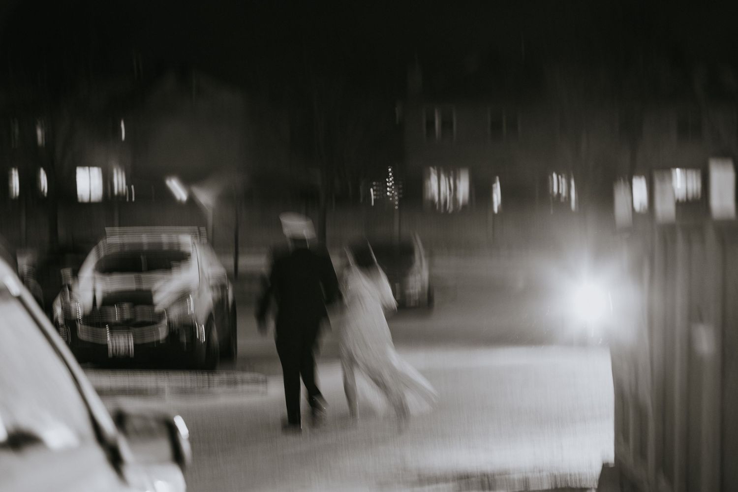 A blurred black and white nighttime street scene with silhouettes walking between parked cars.