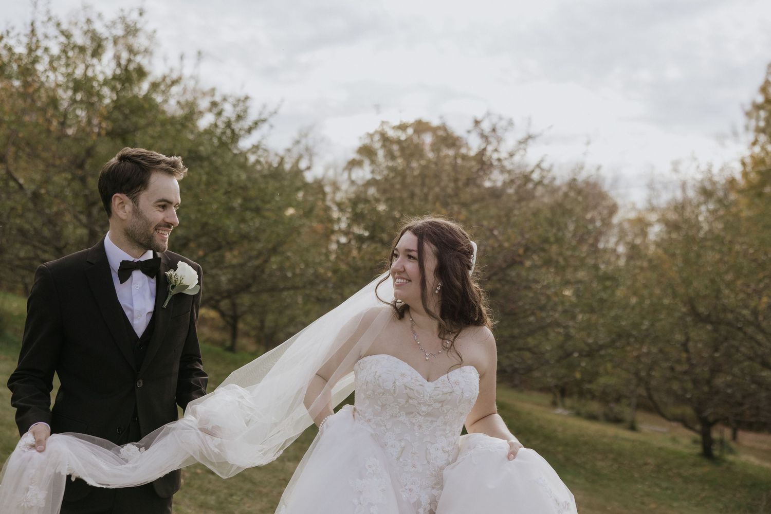 A romantic wedding couple embraces in a scenic autumn orchard with flowing veil and elegant attire.