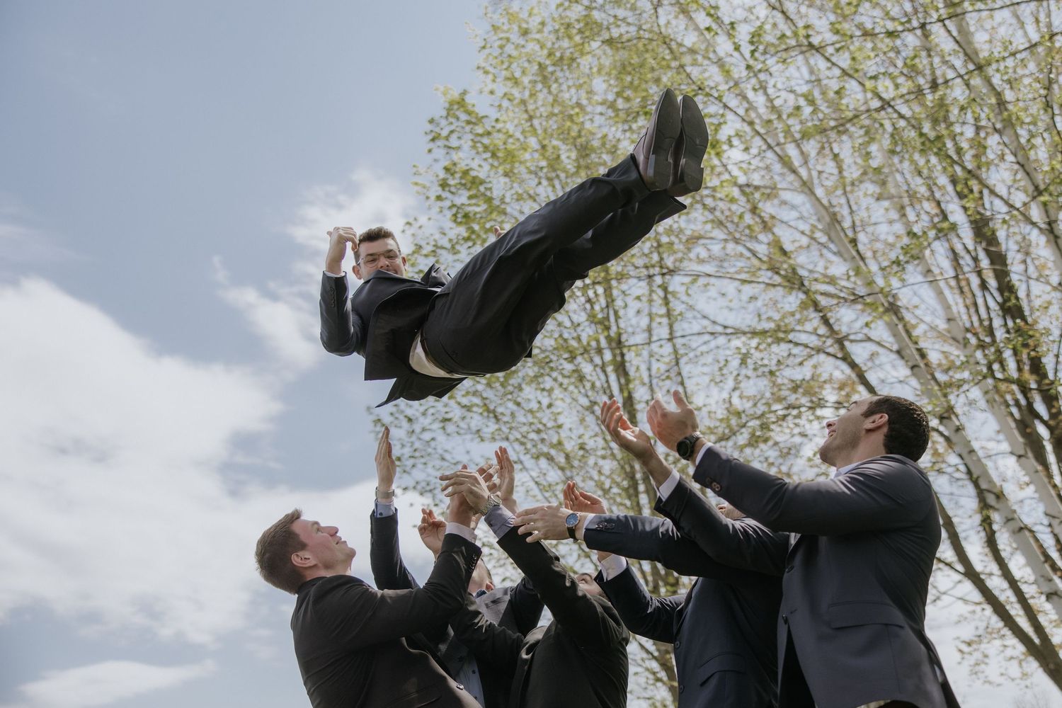 A group of people in dark suits toss someone up into the air during a celebratory moment outdoors.