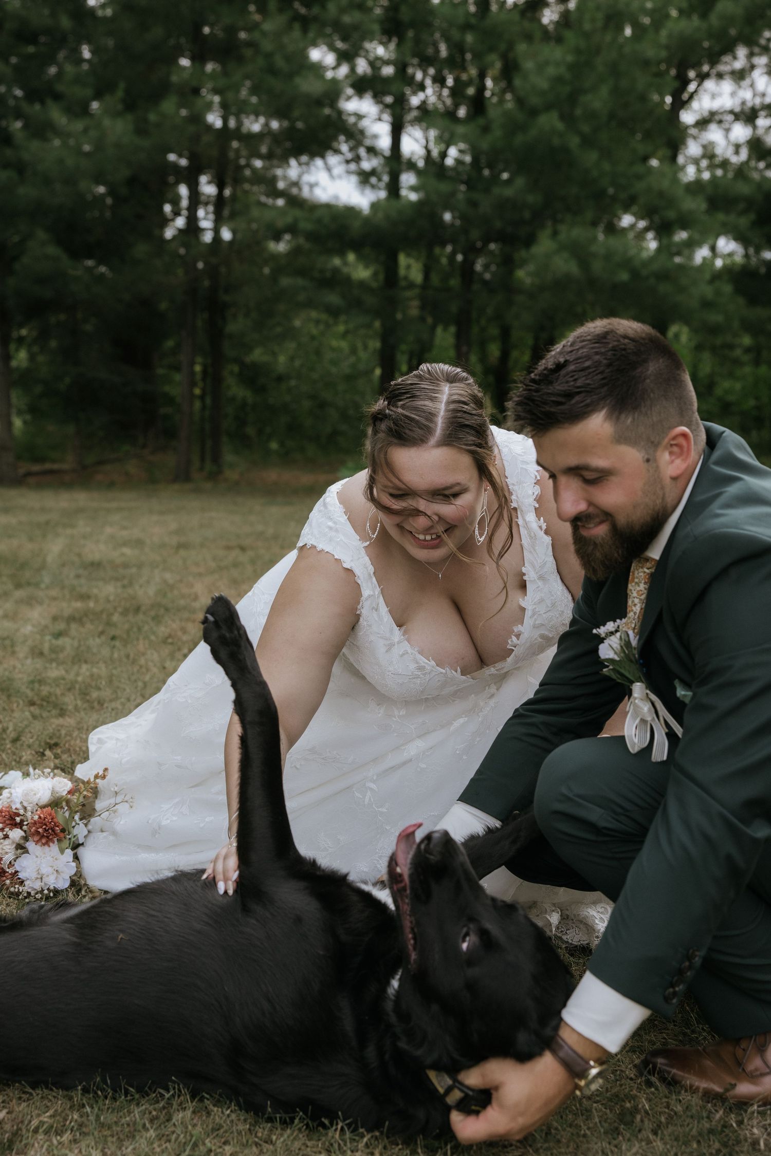 A newlywed couple in wedding attire plays with their black dog on grassy ground during an outdoor photo session.