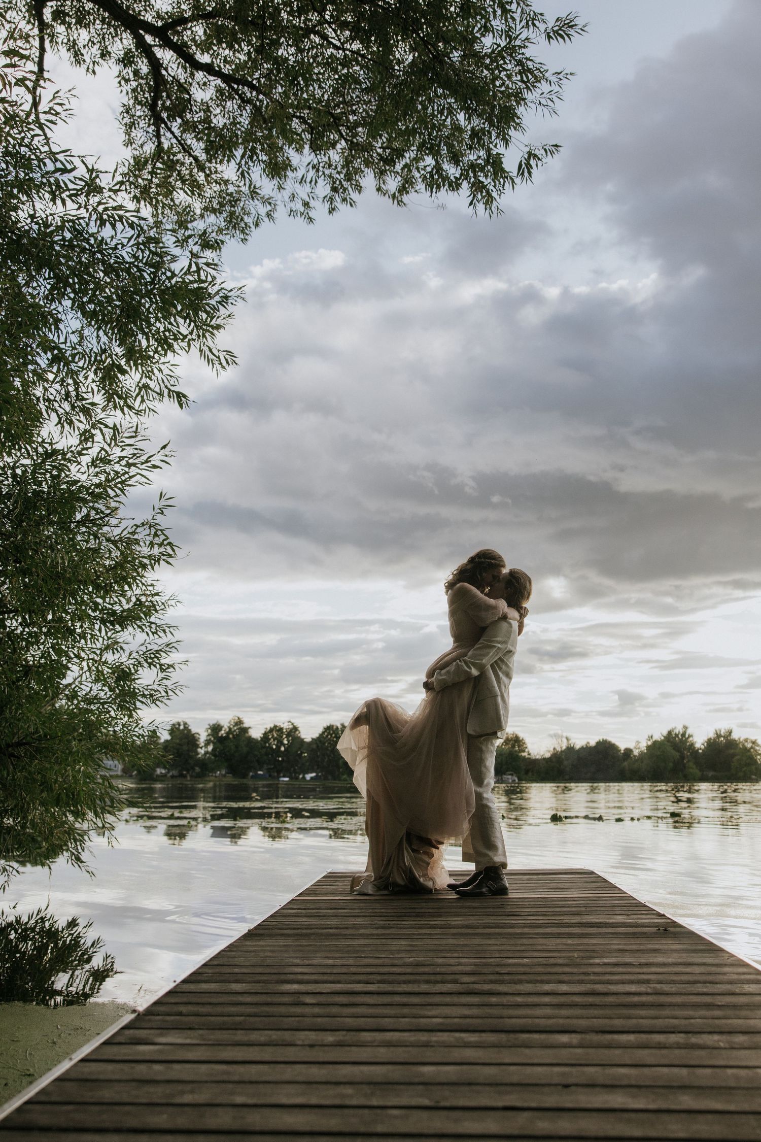 Dog sitting peacefully on a wooden dock overlooking a serene lake under cloudy skies.