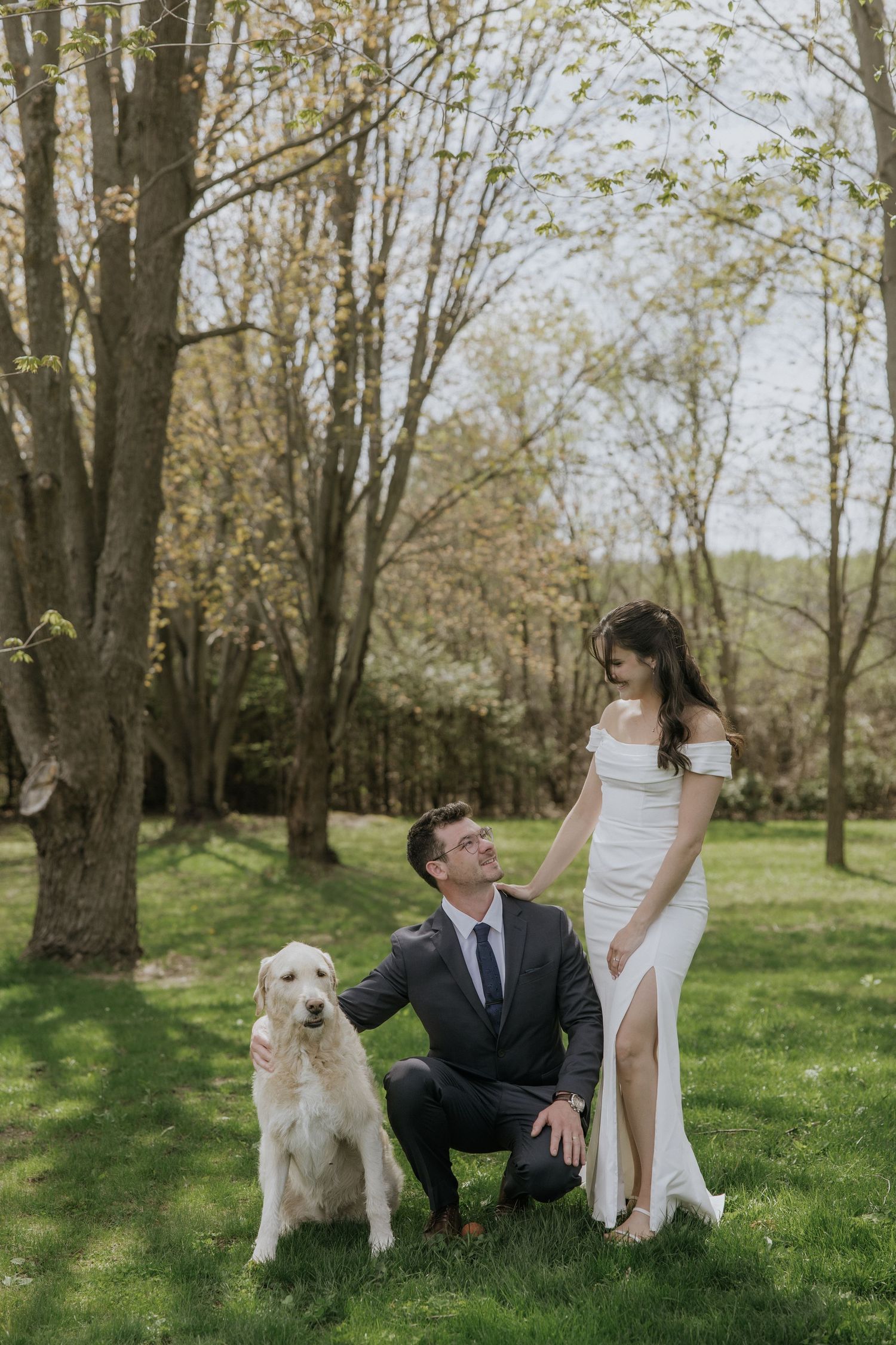 A couple poses with their golden retriever in a sun-dappled spring garden during their wedding photoshoot.