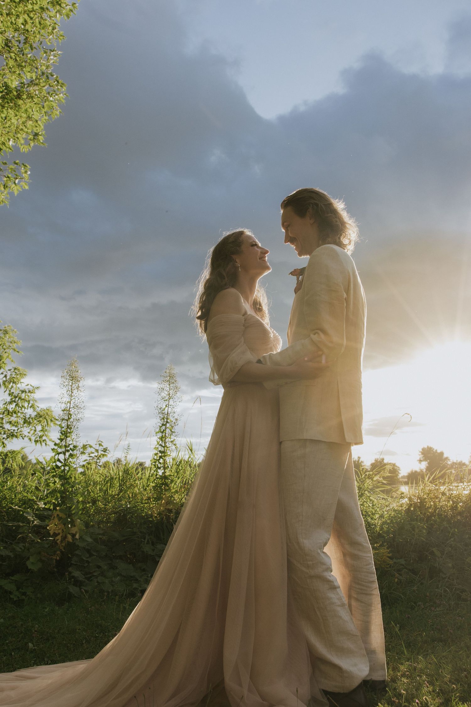 Two figures in flowing cream-colored outfits embrace against a dramatic sunset sky with foliage in background.