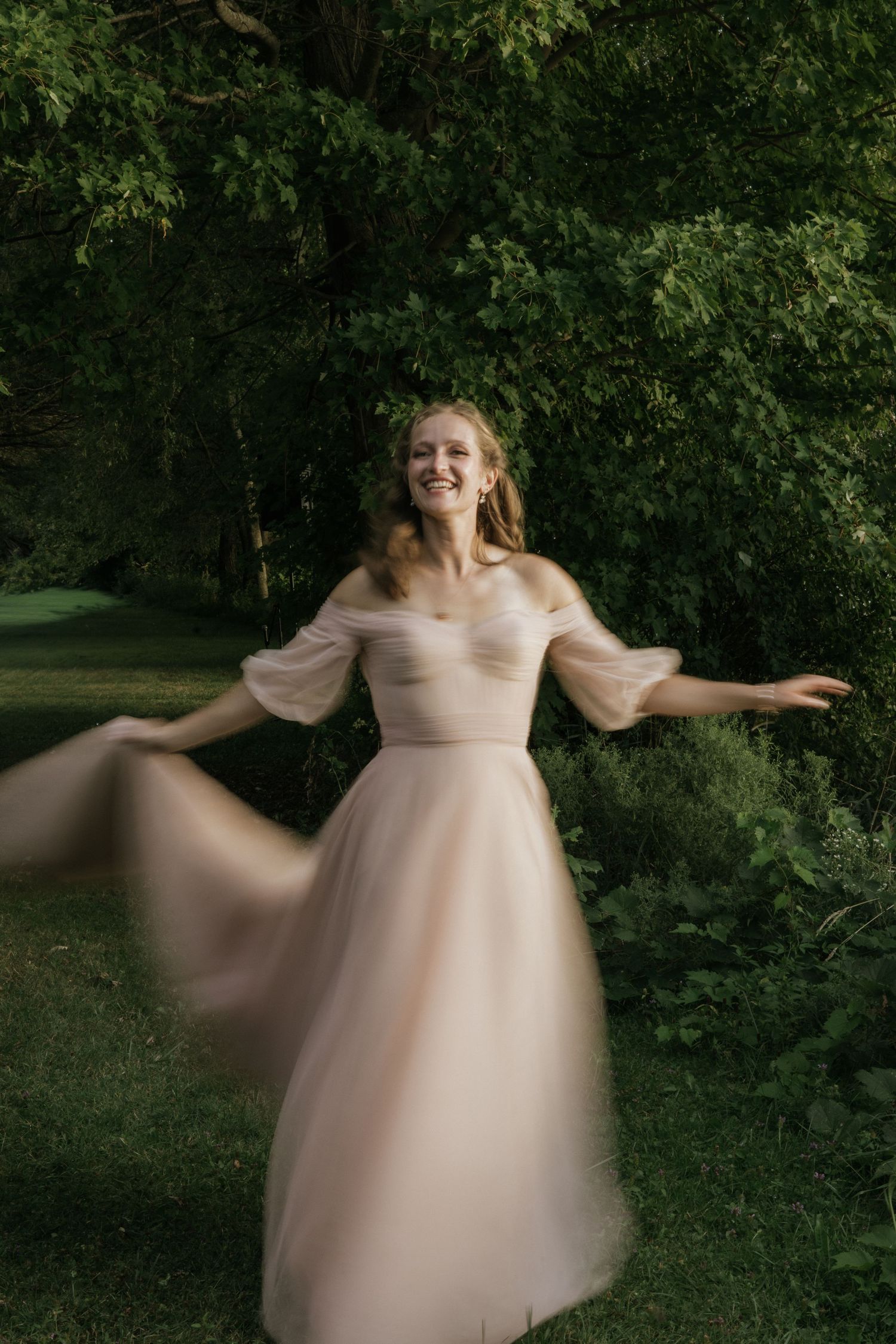 A person in a flowing blush pink off-shoulder gown twirls joyfully in a lush garden setting.