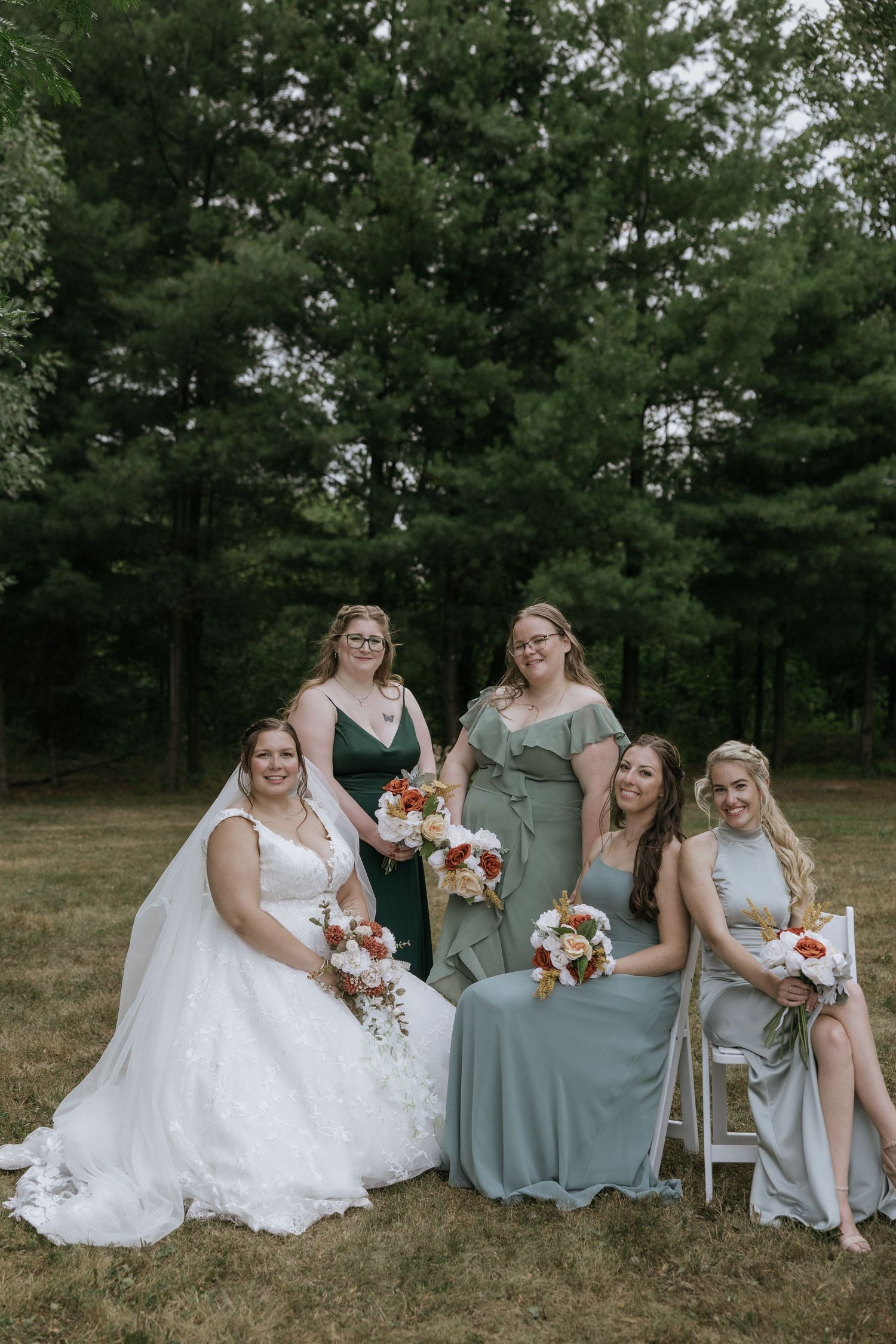 A bridal party in sage green dresses sits together on the grass for outdoor wedding photos with pine trees in background.